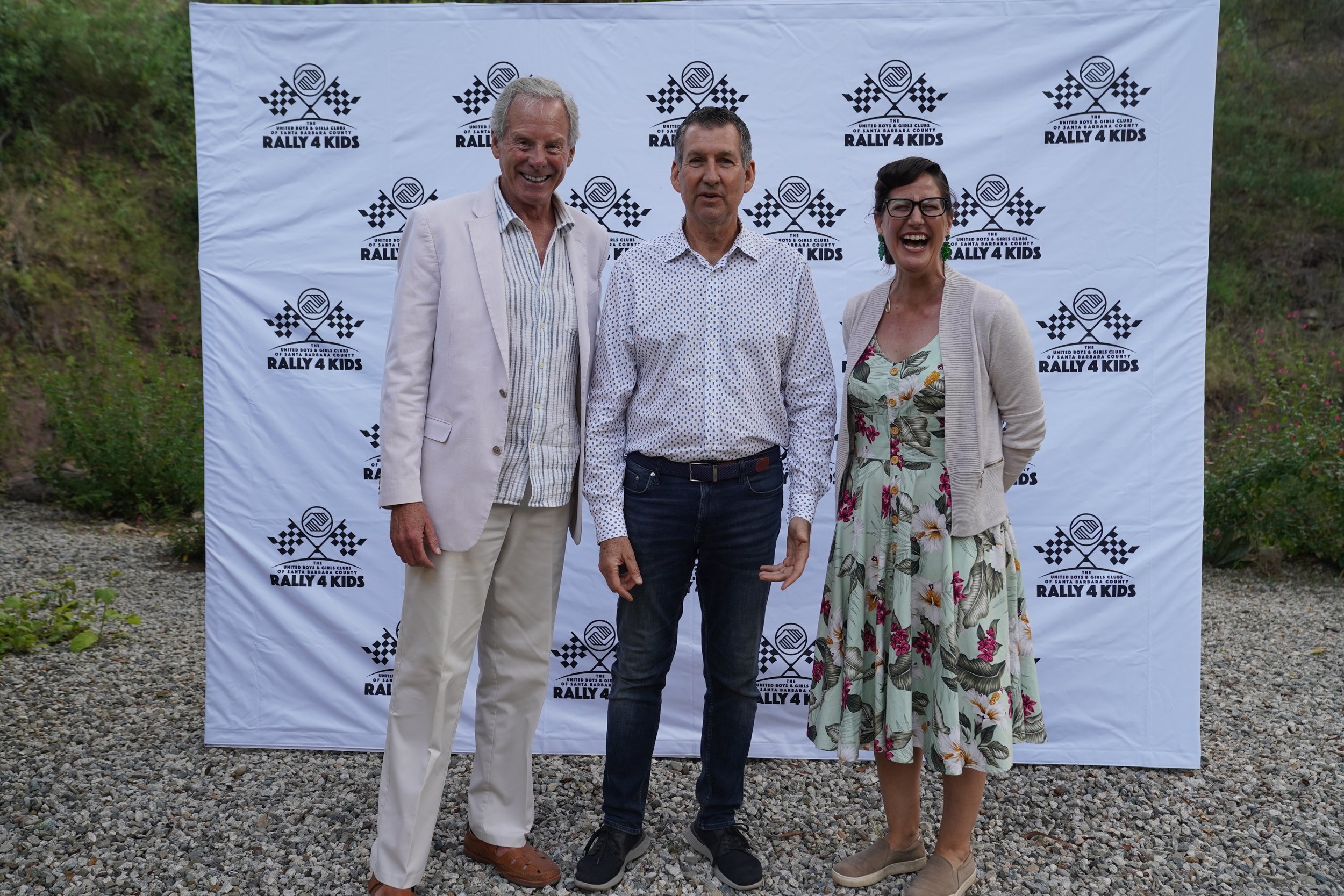 Three people standing outdoors in front of a Rally 4 Kids backdrop. They are smiling and posing for the photo, dressed in semi-formal attire.