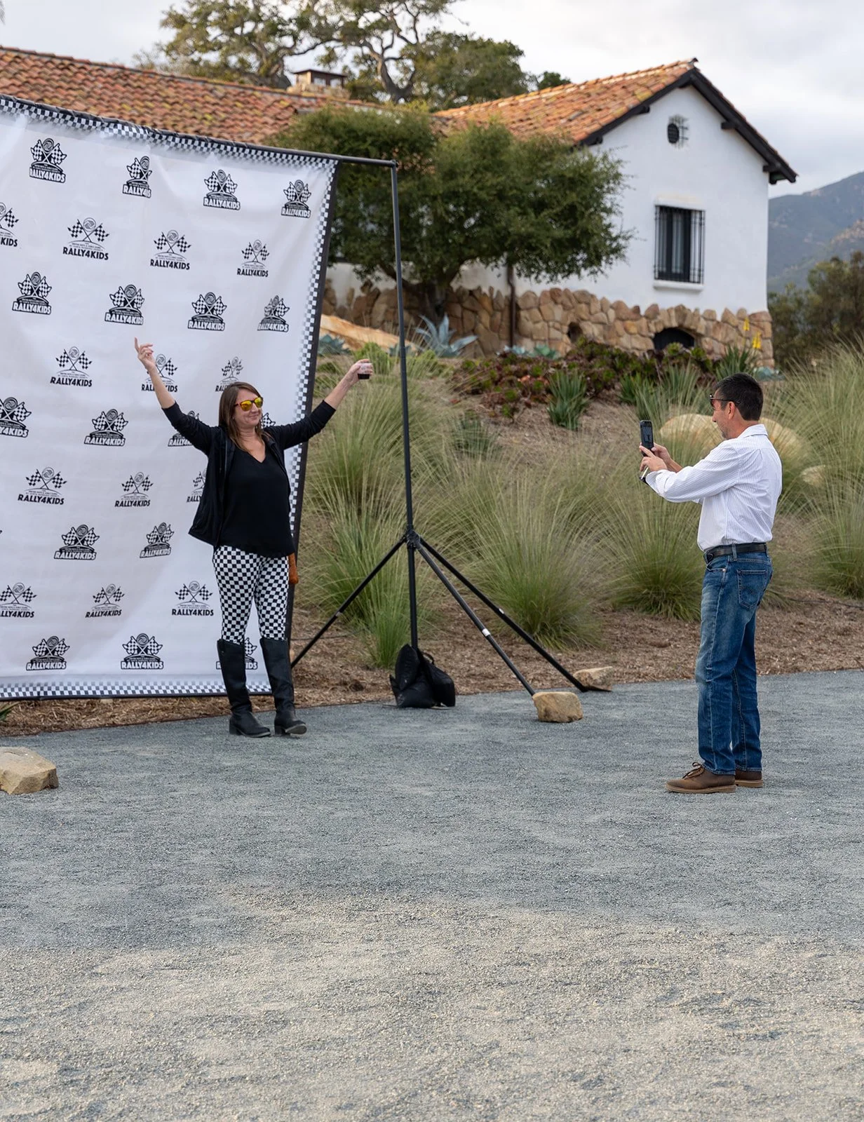 Woman posing with arms raised in front of a Rally4Kids backdrop, man taking her photo with a phone, outdoor setting with house and plants in background.