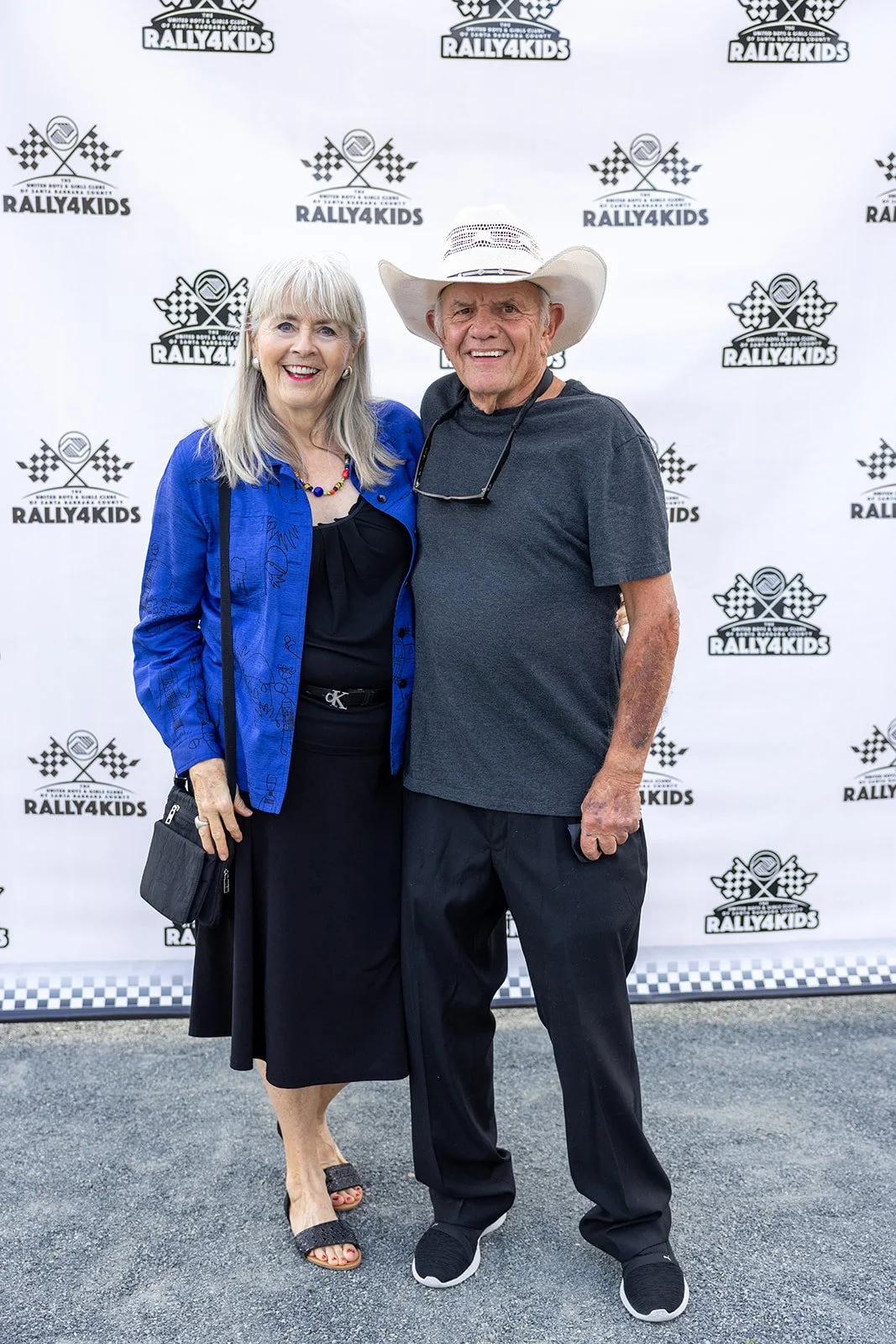 Two smiling adults, a woman with long gray hair wearing a black dress and a blue jacket, and a man with a white cowboy hat wearing a gray t-shirt, standing in front of a backdrop with the Rally4Kids logo, at a rally event.