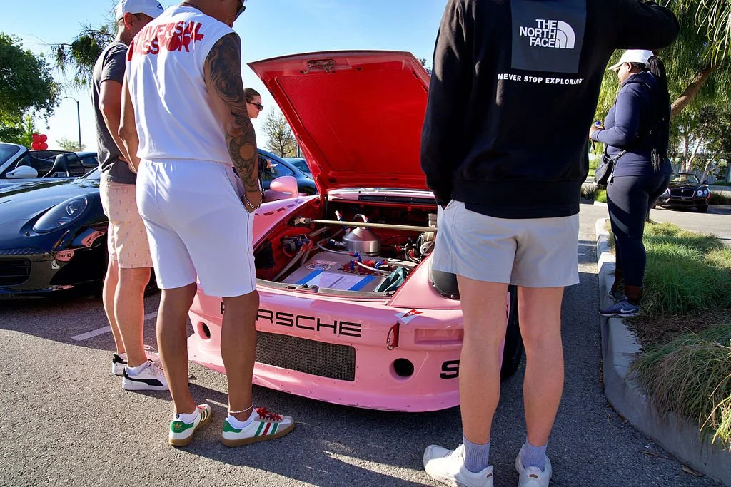 A group of people gathered around a pink Porsche race car with its hood open, inspecting the engine at an outdoor car show on a sunny day.