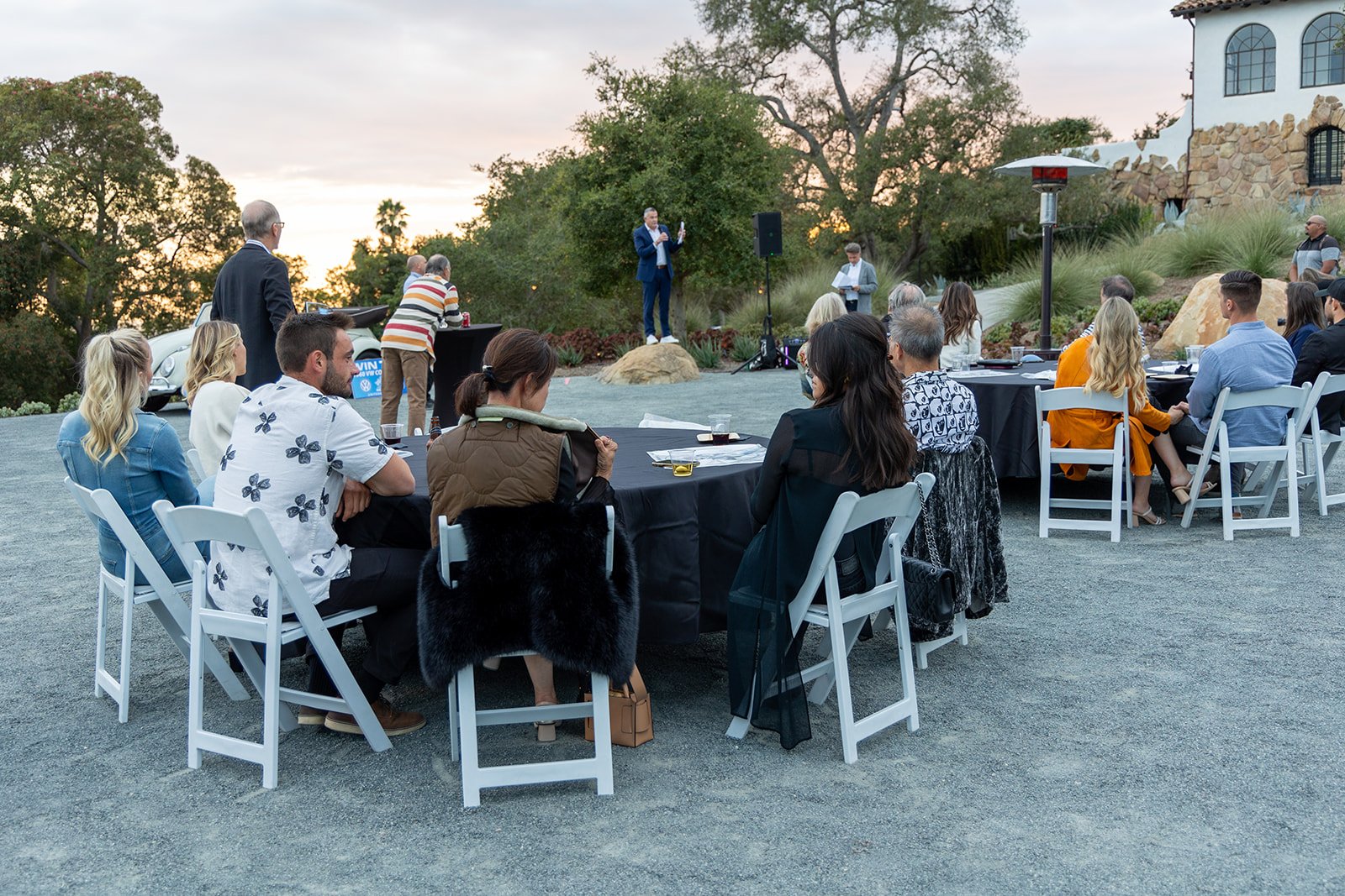 People attending an outdoor event sitting at tables, listening to a speaker on a stage with a scenic background at sunset.
