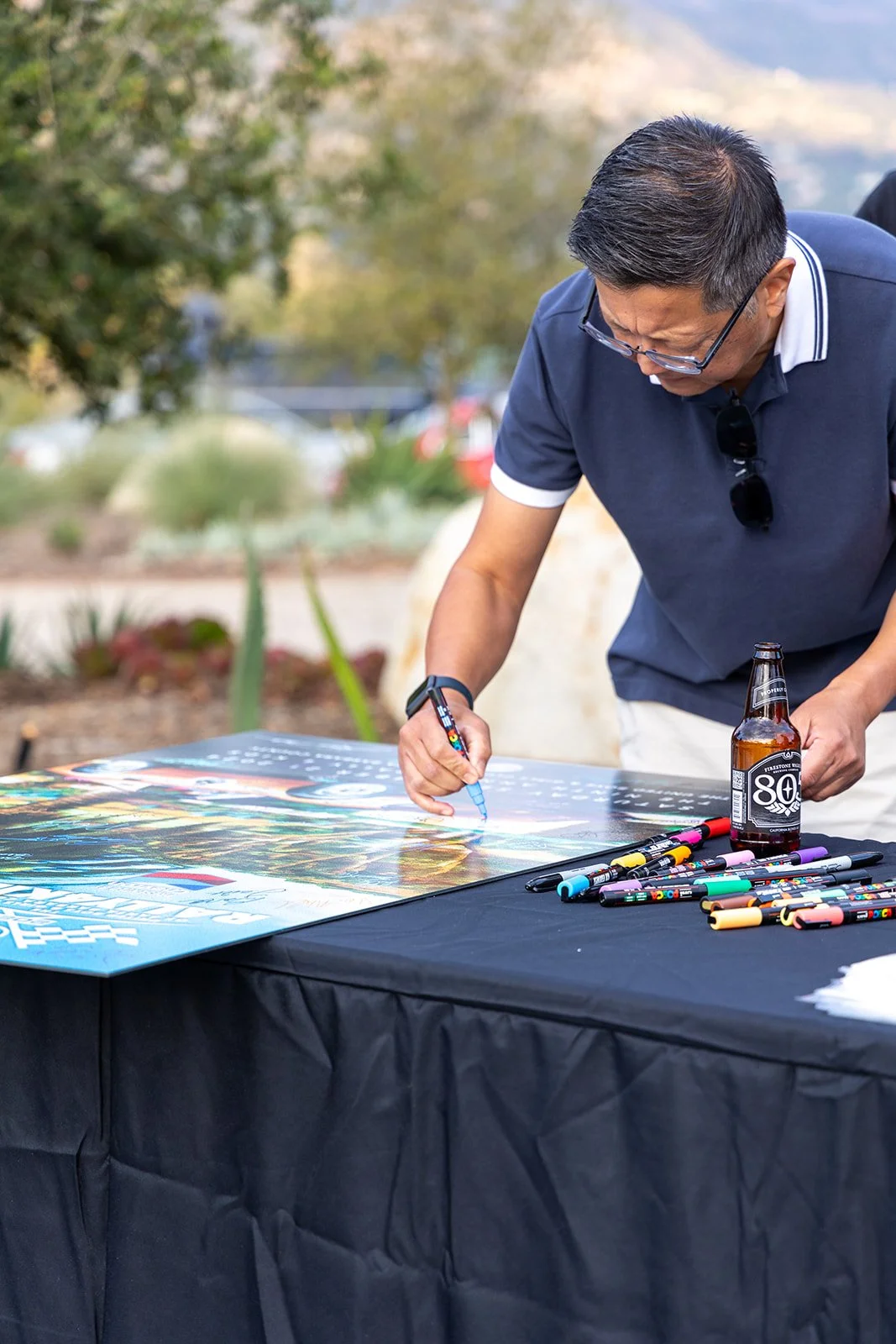 Man with glasses signing a sports poster at an outdoor event, standing at a black table with markers and a beer bottle.