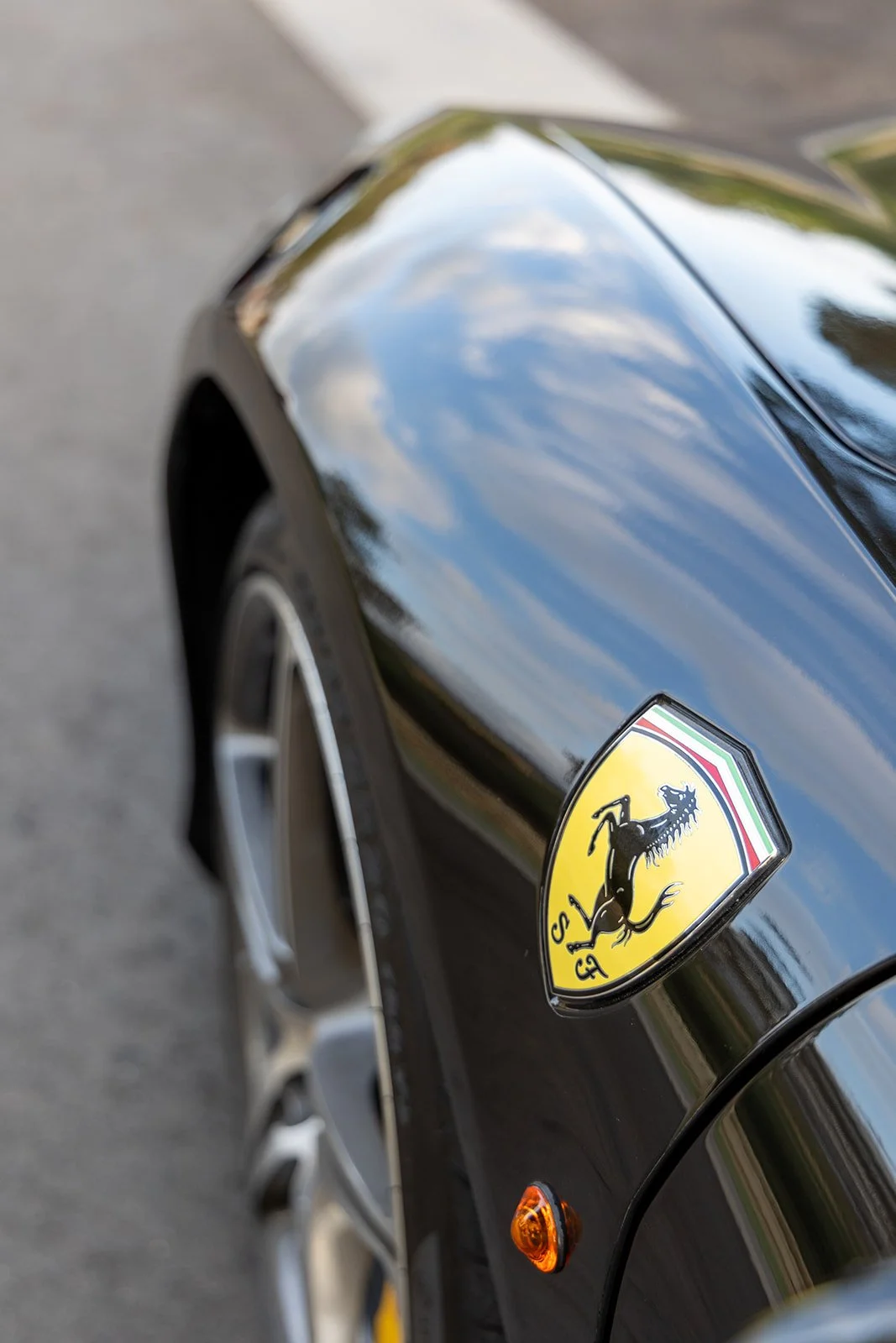 Close-up of a black Ferrari car with reflections of the sky and trees, showing the Ferrari emblem with a black prancing horse on a yellow shield.