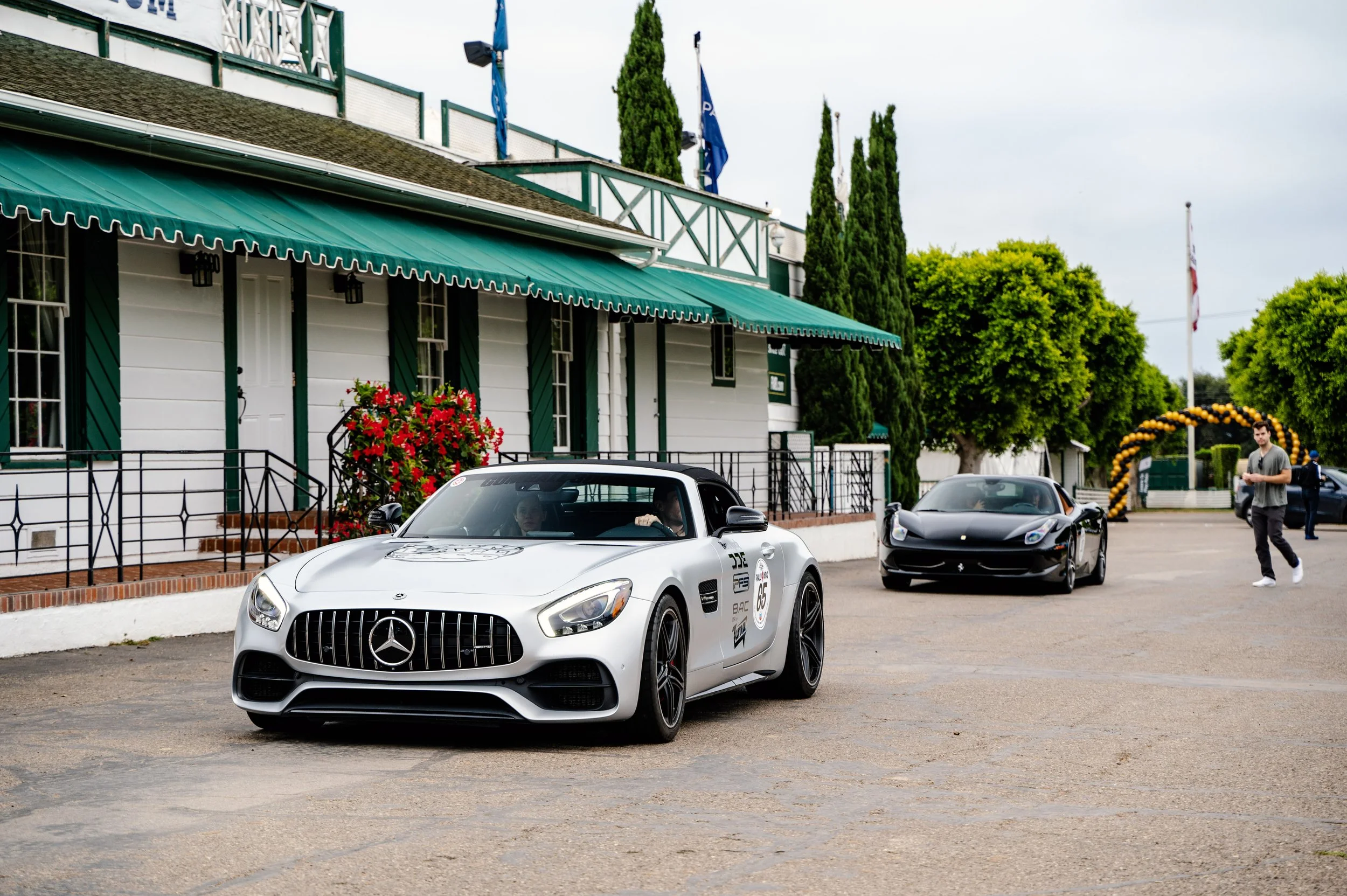 A white Mercedes-Benz and a black Ferrari parked outside a building with a green awning and landscaped trees, with a man walking nearby.