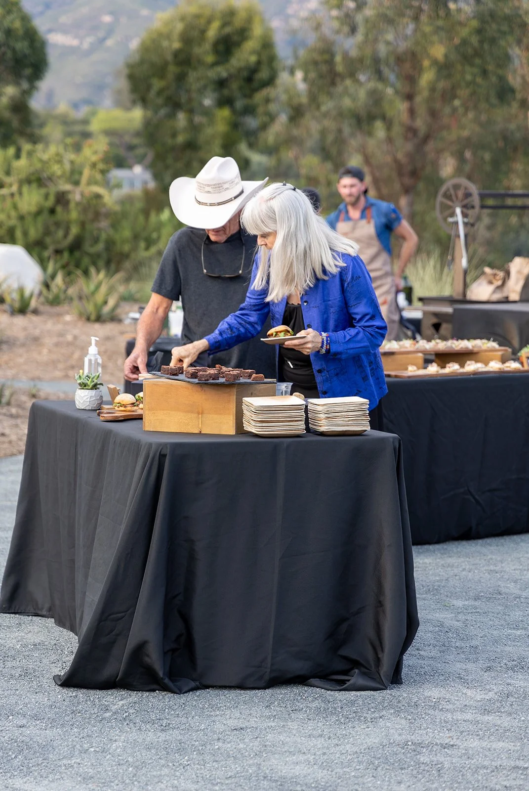 People serving food at an outdoor event with black tablecloths and a variety of dishes, including brownies, on the tables, in a natural setting with trees and mountains in the background.