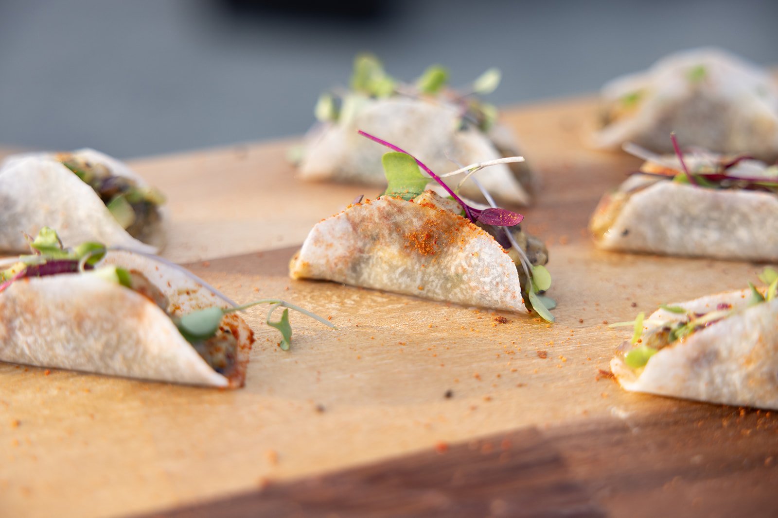 Close-up of several small wrapped tacos with microgreens on a wooden surface, with one taco slightly more visible and unwrapped, showing vegetables inside.