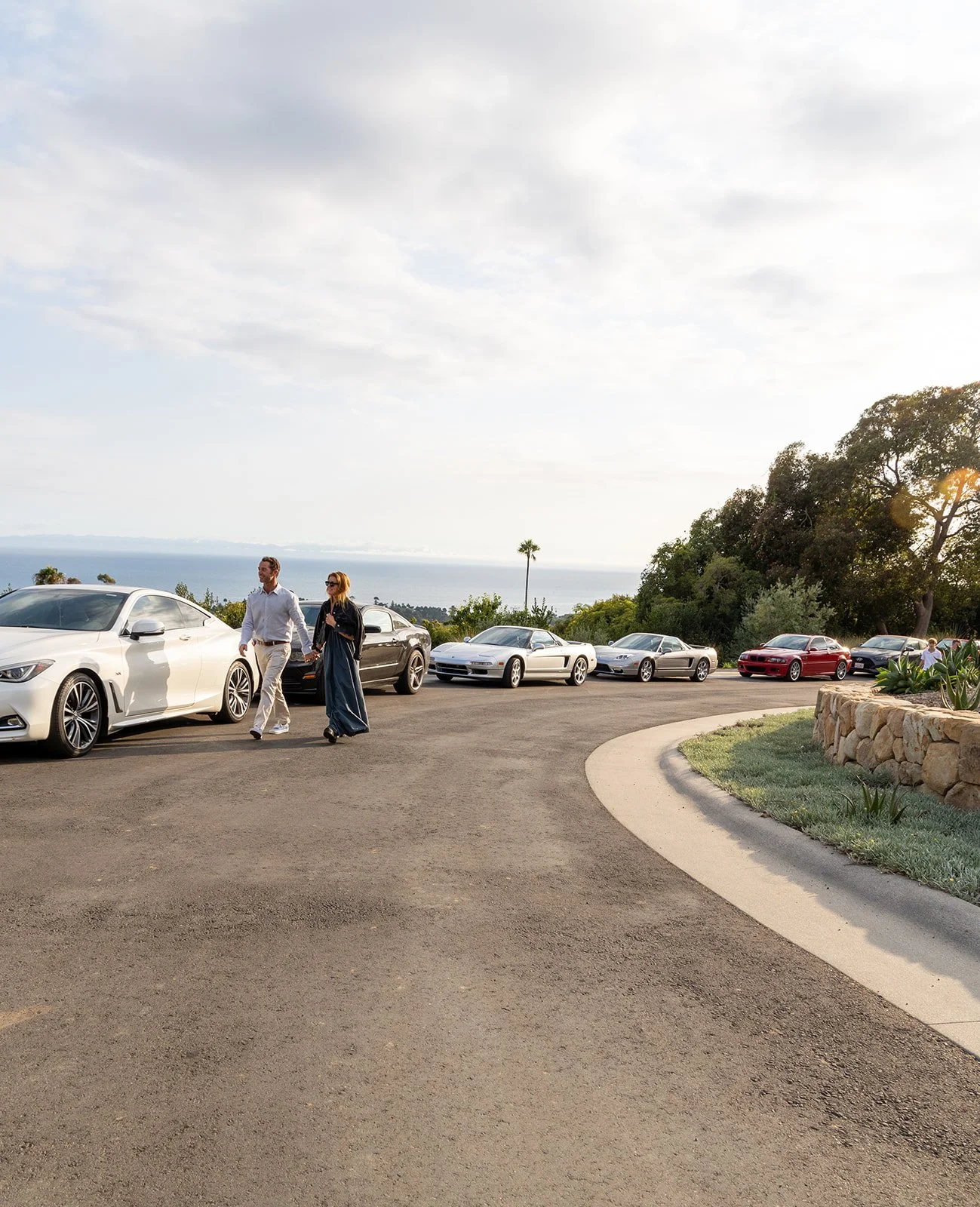 People walking past luxury cars parked on a scenic hillside with greenery, stone wall, and ocean view in the background.
