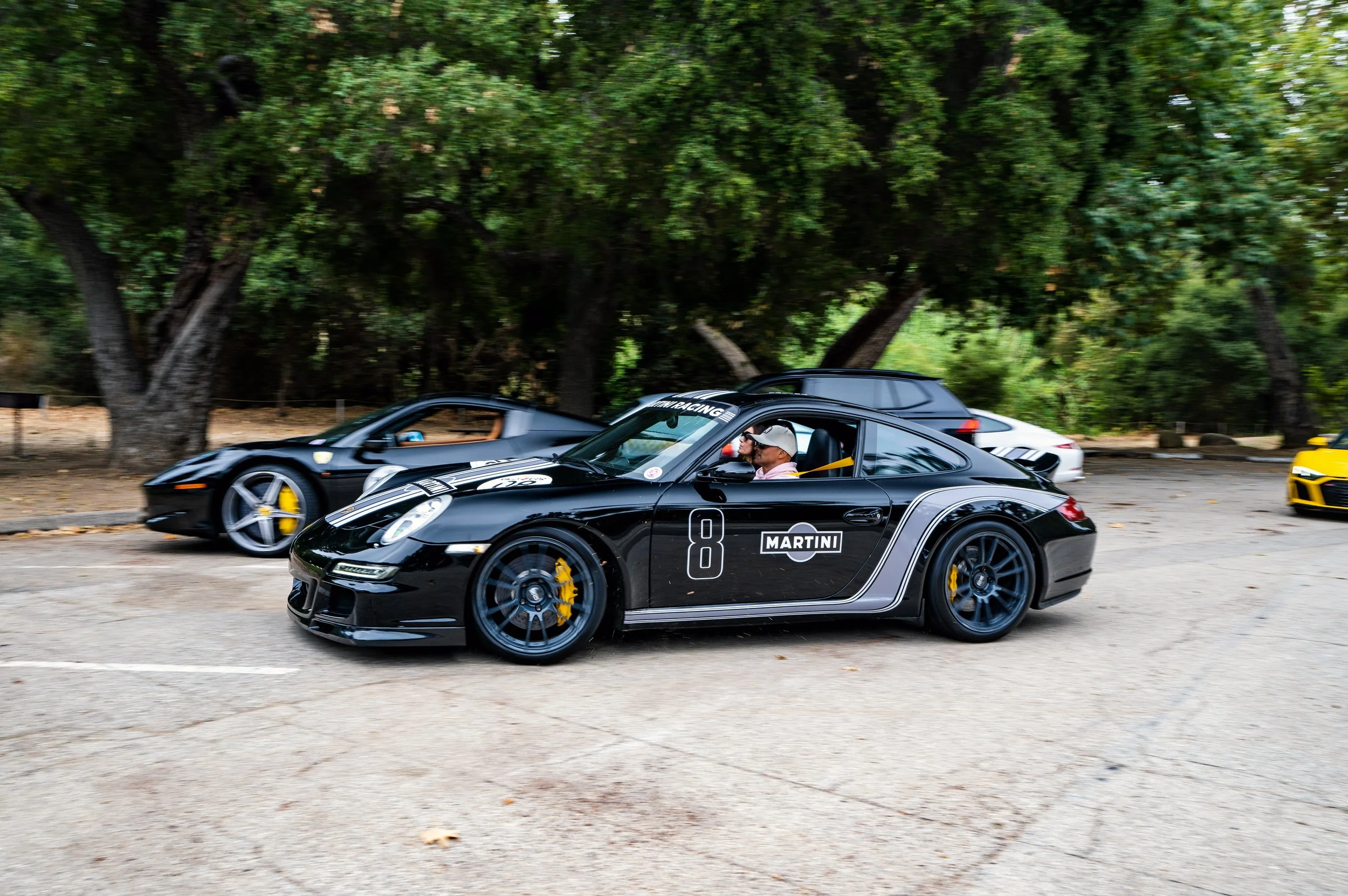 Multiple luxury sports cars, including a black Porsche with racing stripes and a Martini logo, parked on a tree-lined street.