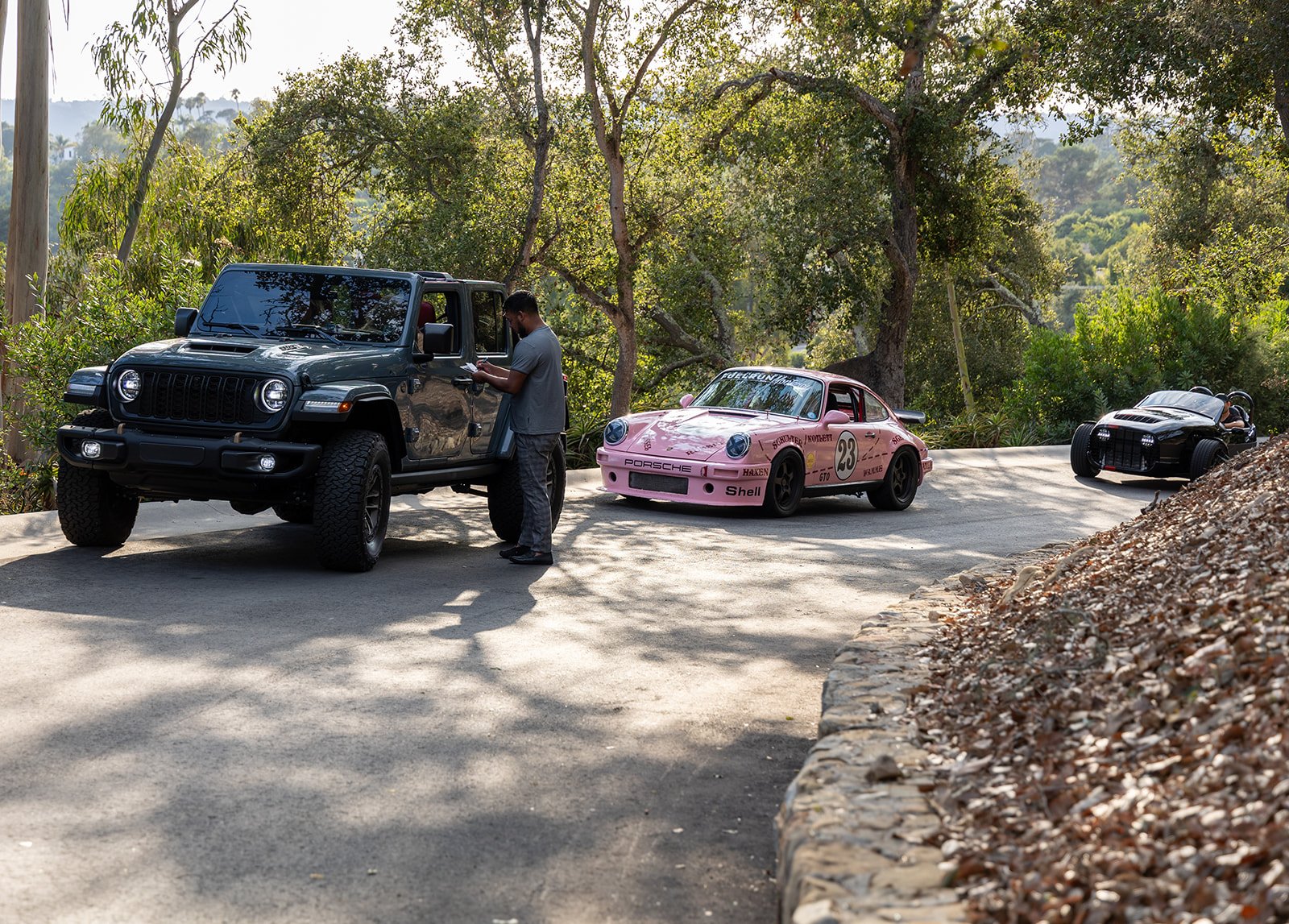 A man standing beside a black Jeep, while two race cars, a pink Porsche and a black race car, are parked on a dirt road surrounded by trees.