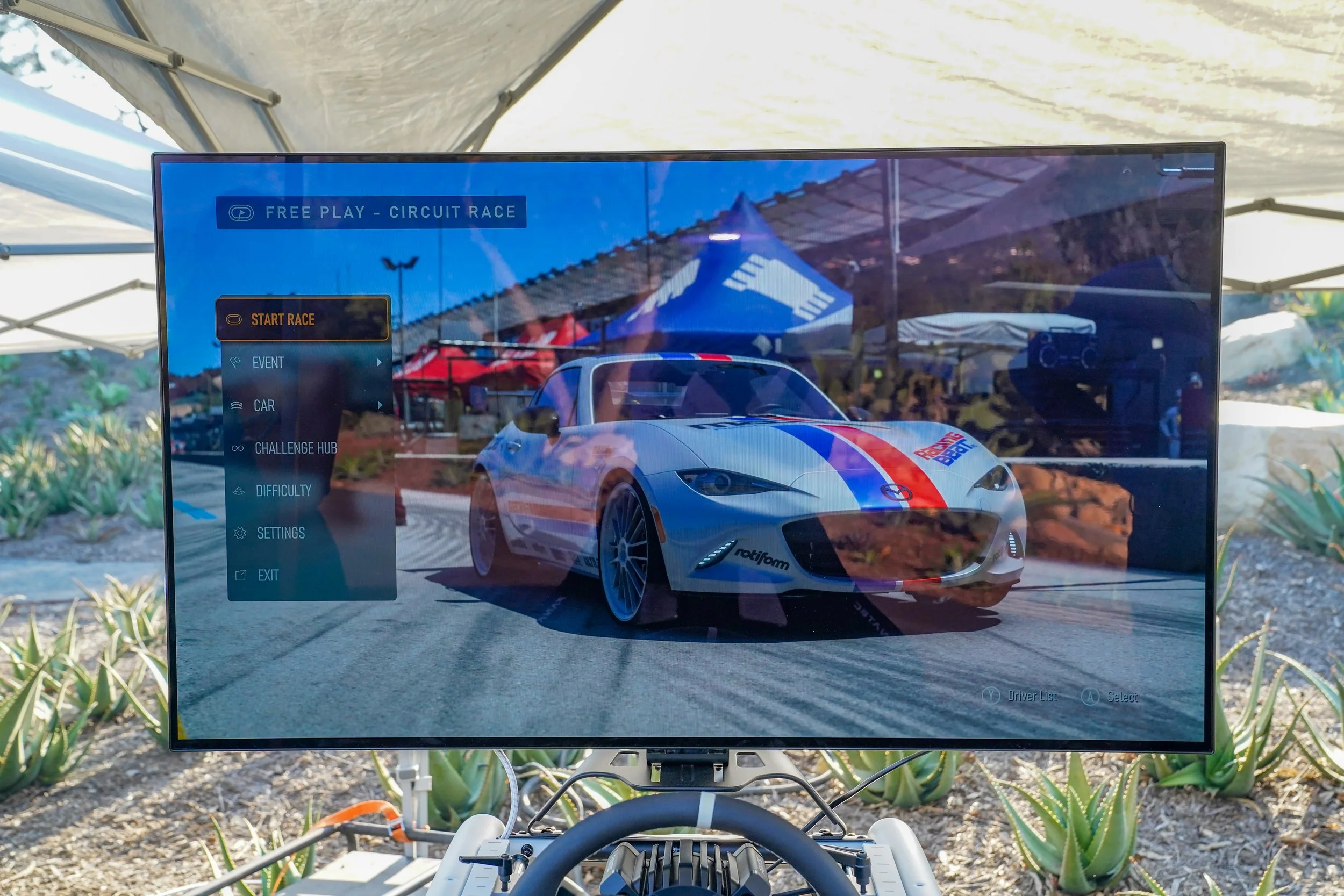 A racing video game displayed on a large screen, showing a sports car at a race track with options menu on the left side.