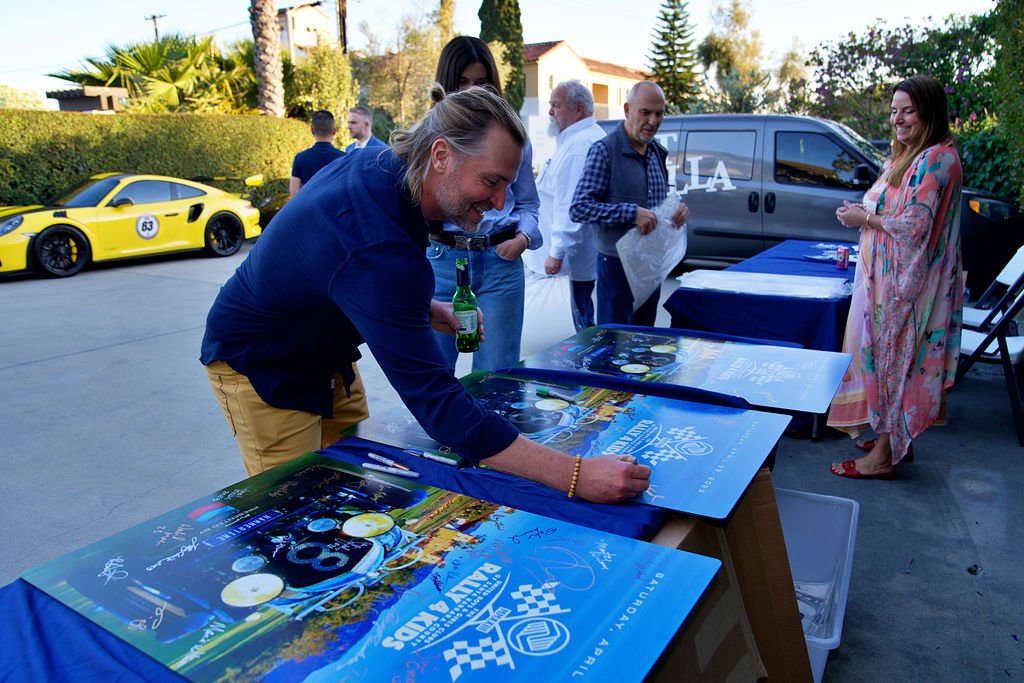 Man signing posters at an outdoor event with a yellow sports car in the background.