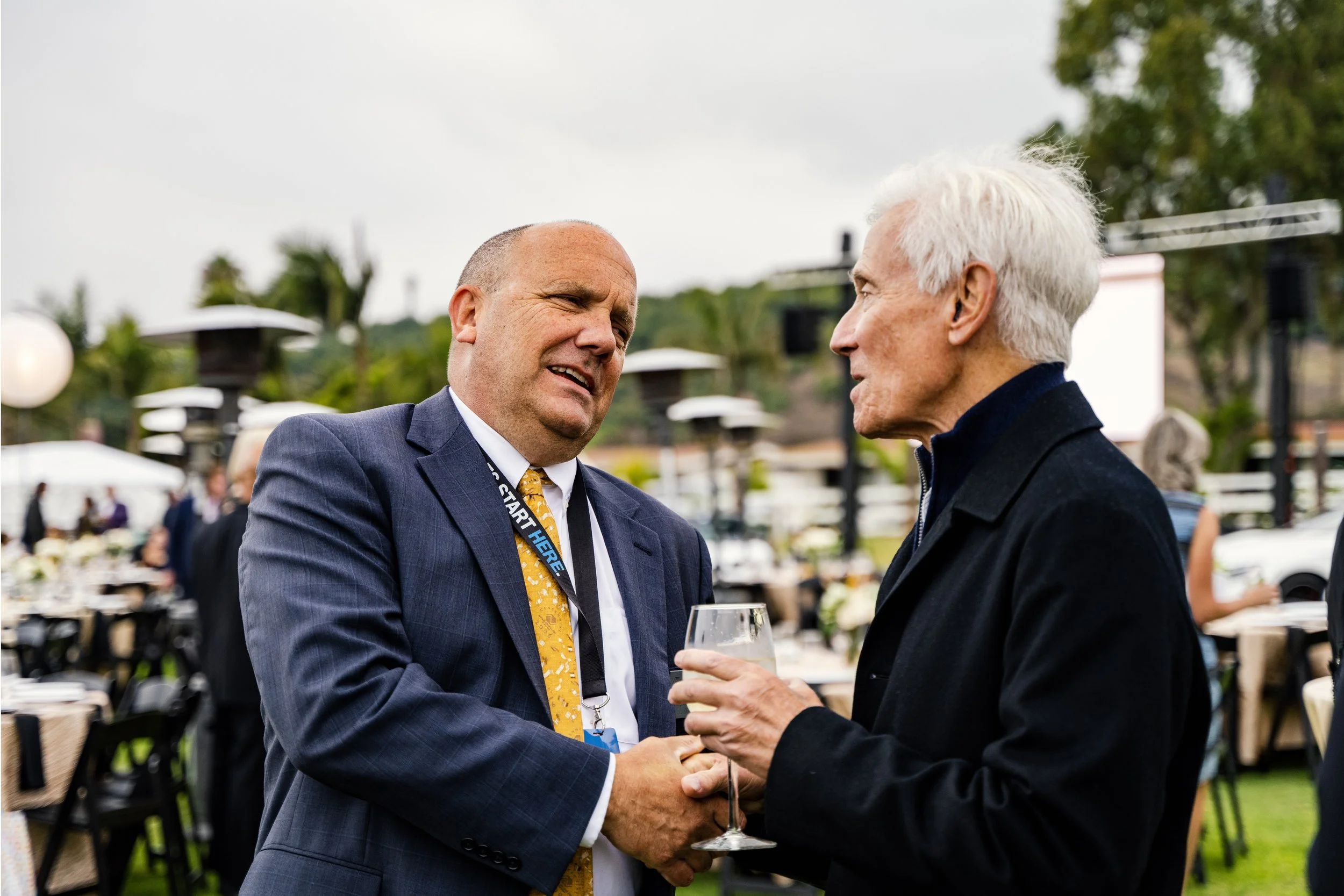 Two men in formal attire engaged in conversation at an outdoor event, with one holding a glass of wine.