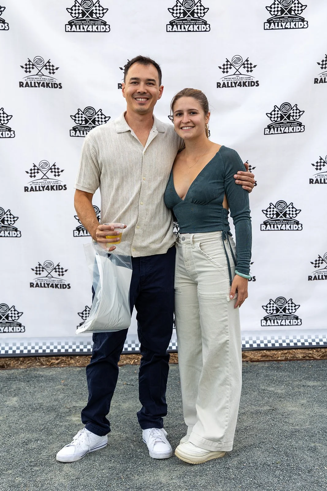 A man and a woman standing close together, smiling, with a backdrop that says 'Rally4Kids' and features checkered flags. The man is holding a drink and a plastic bag, and the woman has her arm around his waist.