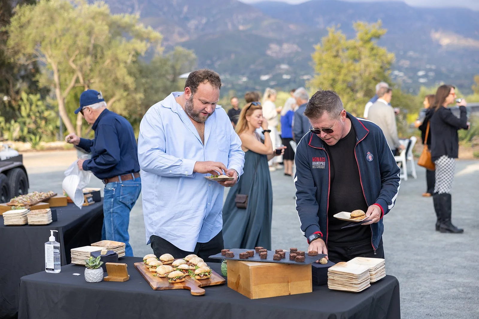 People at an outdoor event enjoying food, with a table of sandwiches and brownies, and a scenic mountain background.