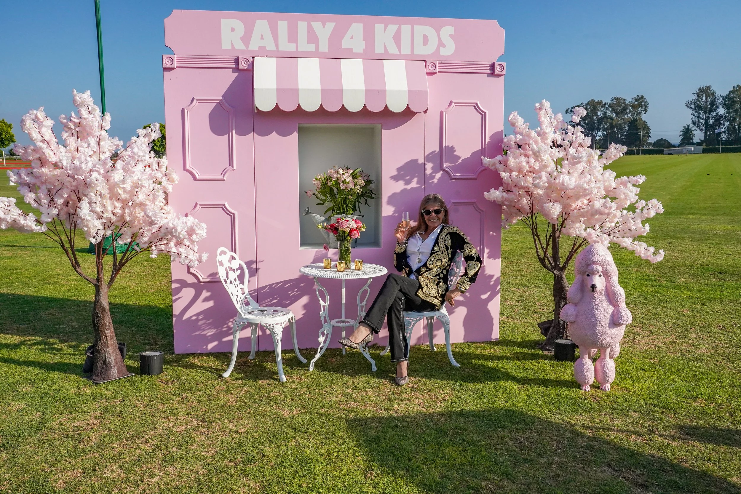 Woman smiling and raising a glass of champagne while sitting at a small white garden table with two chairs in front of a pink backdrop labeled 'RALLY 4 KIDS.' The scene is decorated with pink cherry blossom trees and a pink poodle sculpture, all set 