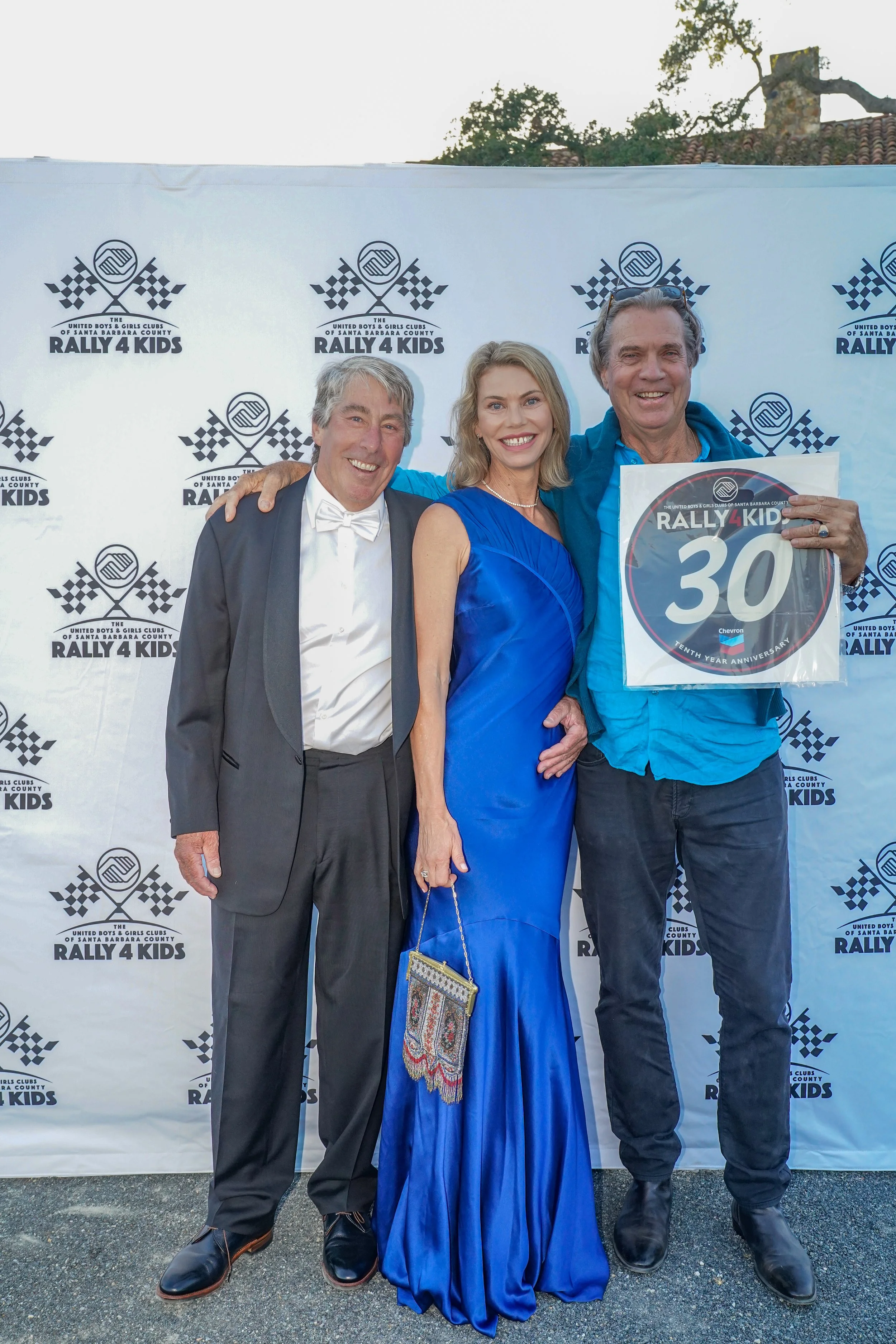 Three people dressed in formal and semi-formal attire pose together at a rally event, with a backdrop displaying the event's logo. The man on the right is holding a sign indicating a 30-year anniversary celebration.