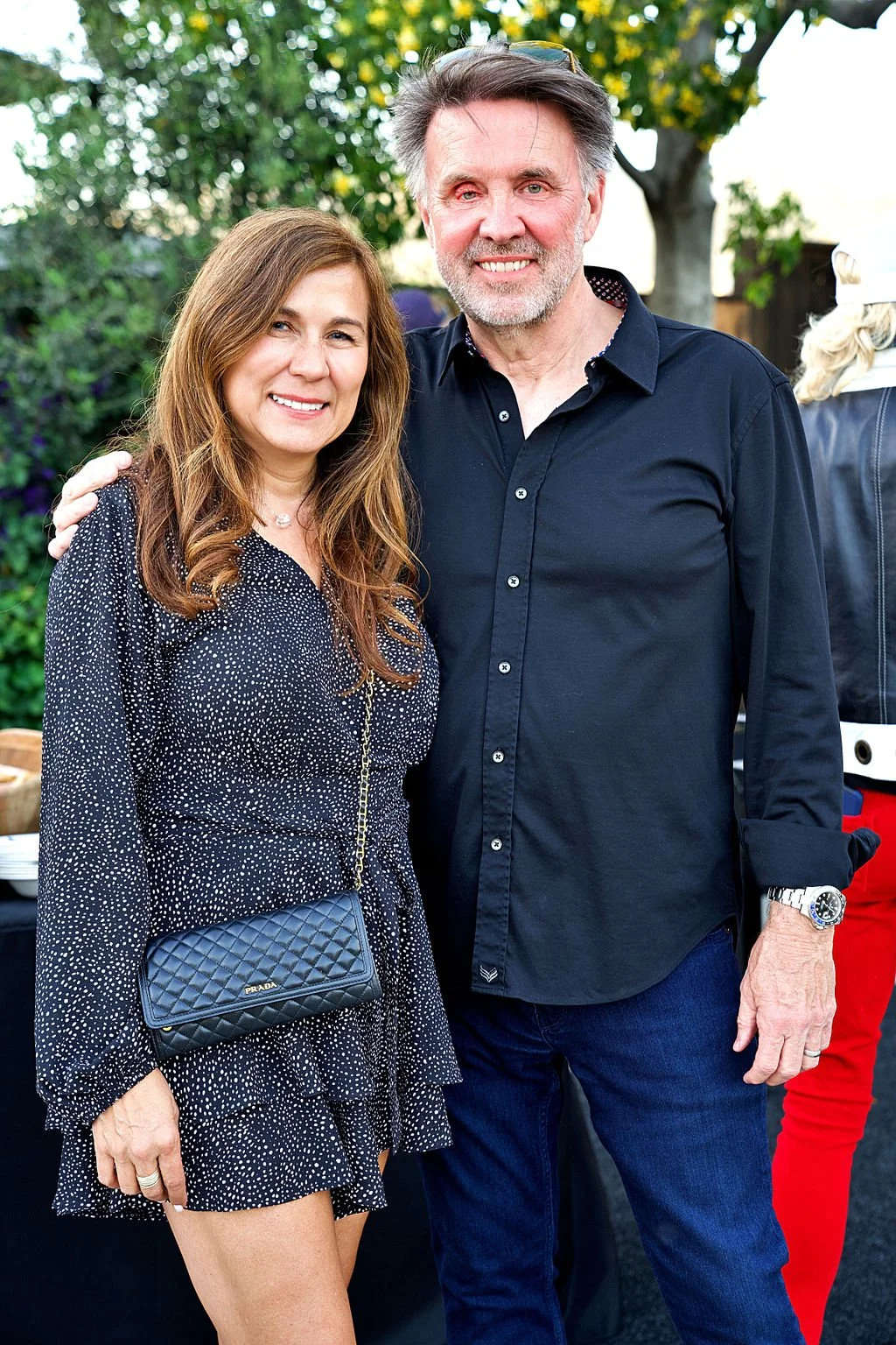 A man and woman standing outdoors, smiling, with trees and a person in the background.