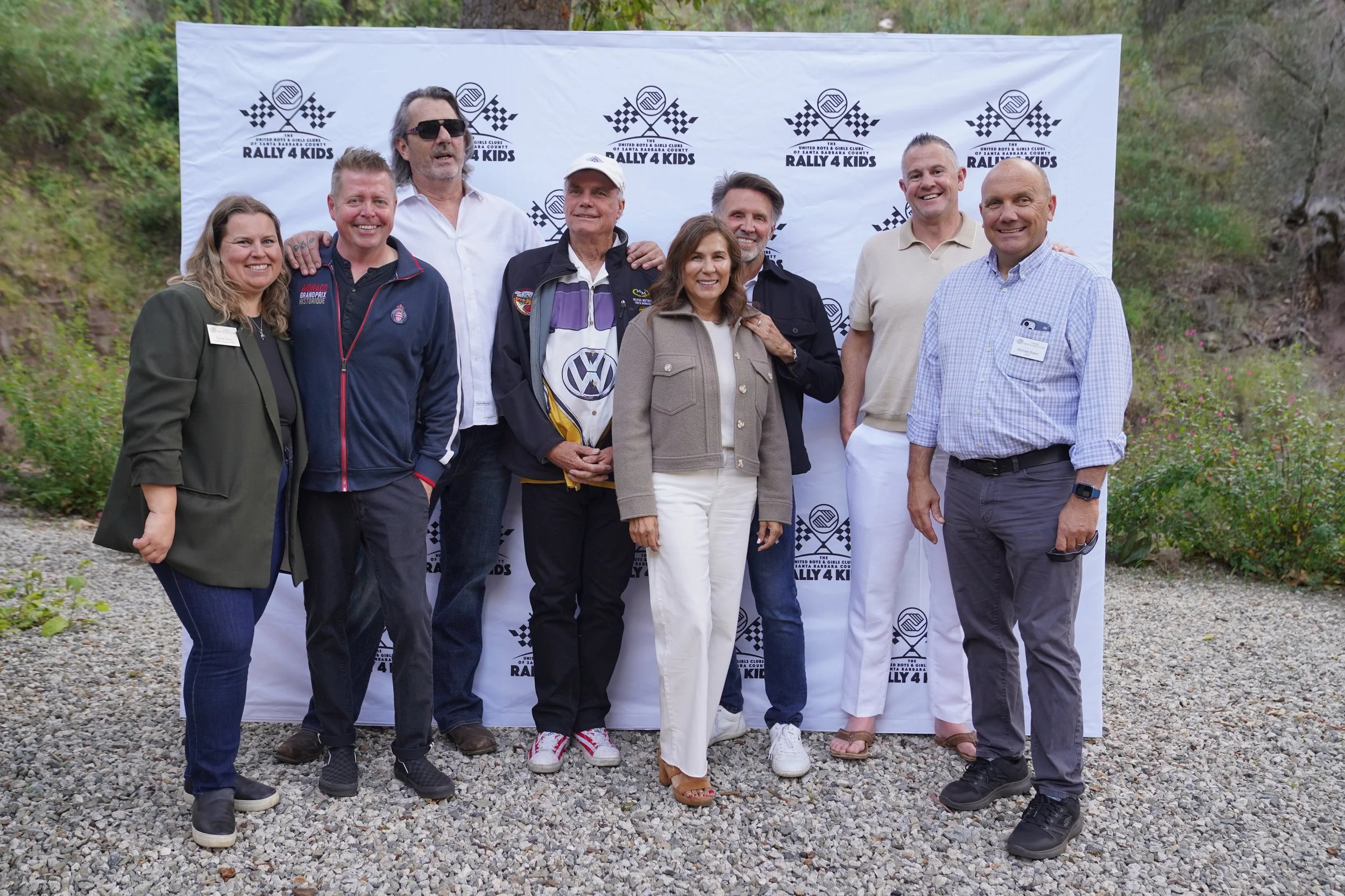 Group of eight people standing outdoors on gravel in front of a white backdrop that reads 'Rally 4 Kids' with racing flags and logos, smiling for a photo.