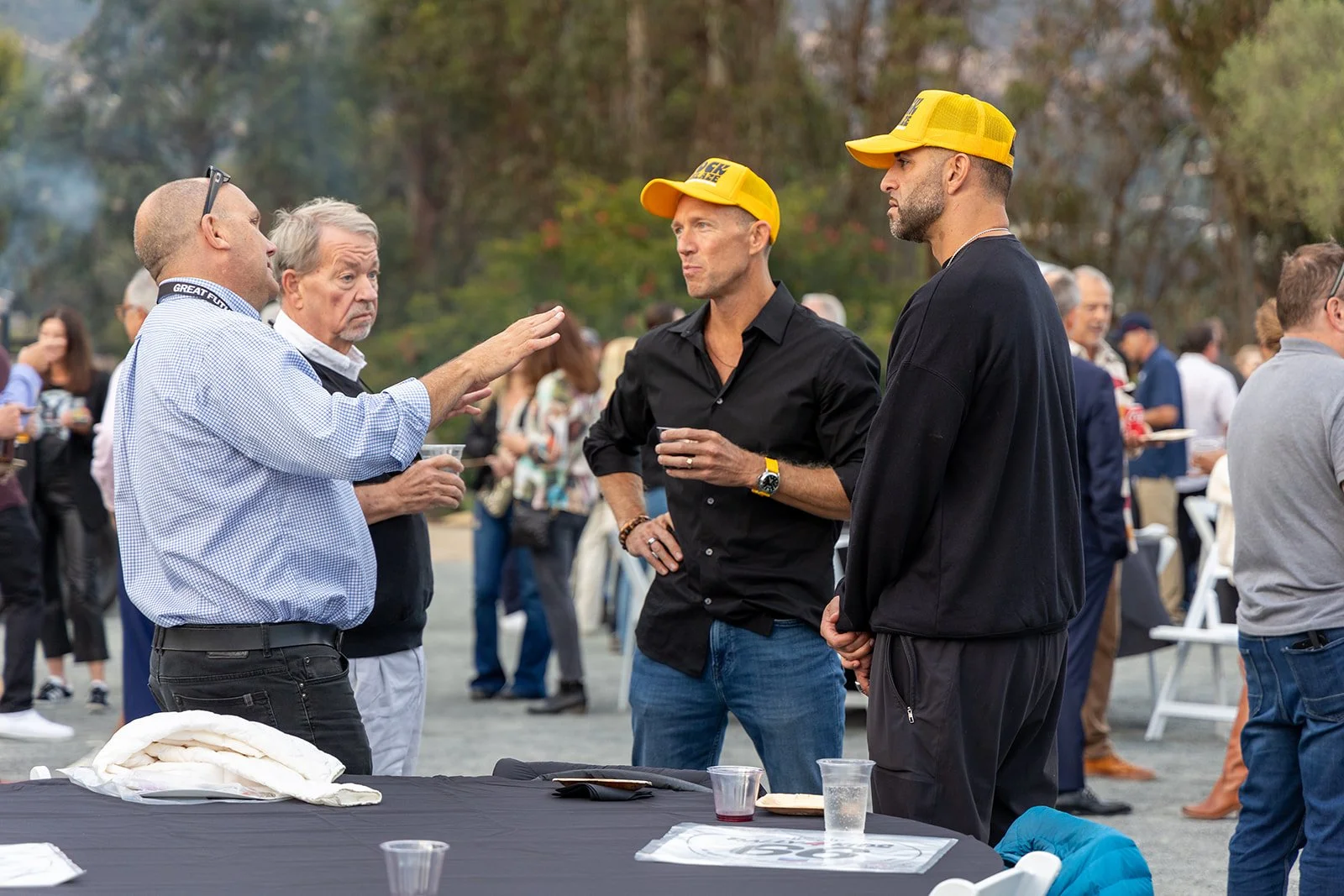 Four men having a conversation at an outdoor event with a crowd in the background. Two of the men are wearing yellow caps, one man in a black shirt with his hand on his hip, and the other in black clothing with hands in his pockets. The man on the le