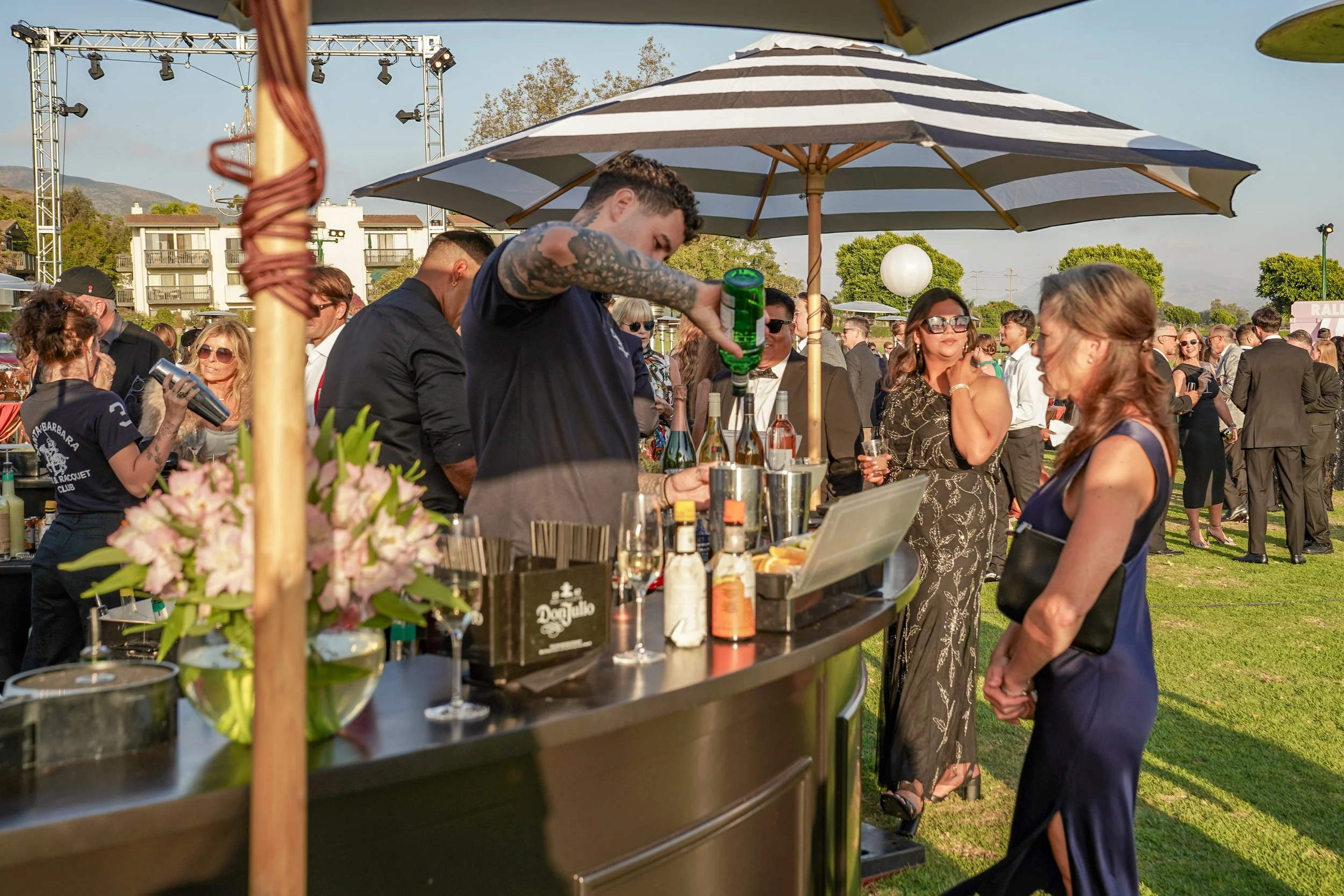 People socializing at an outdoor event with a bar setup, floral arrangements, and a large striped umbrella on a sunny day.