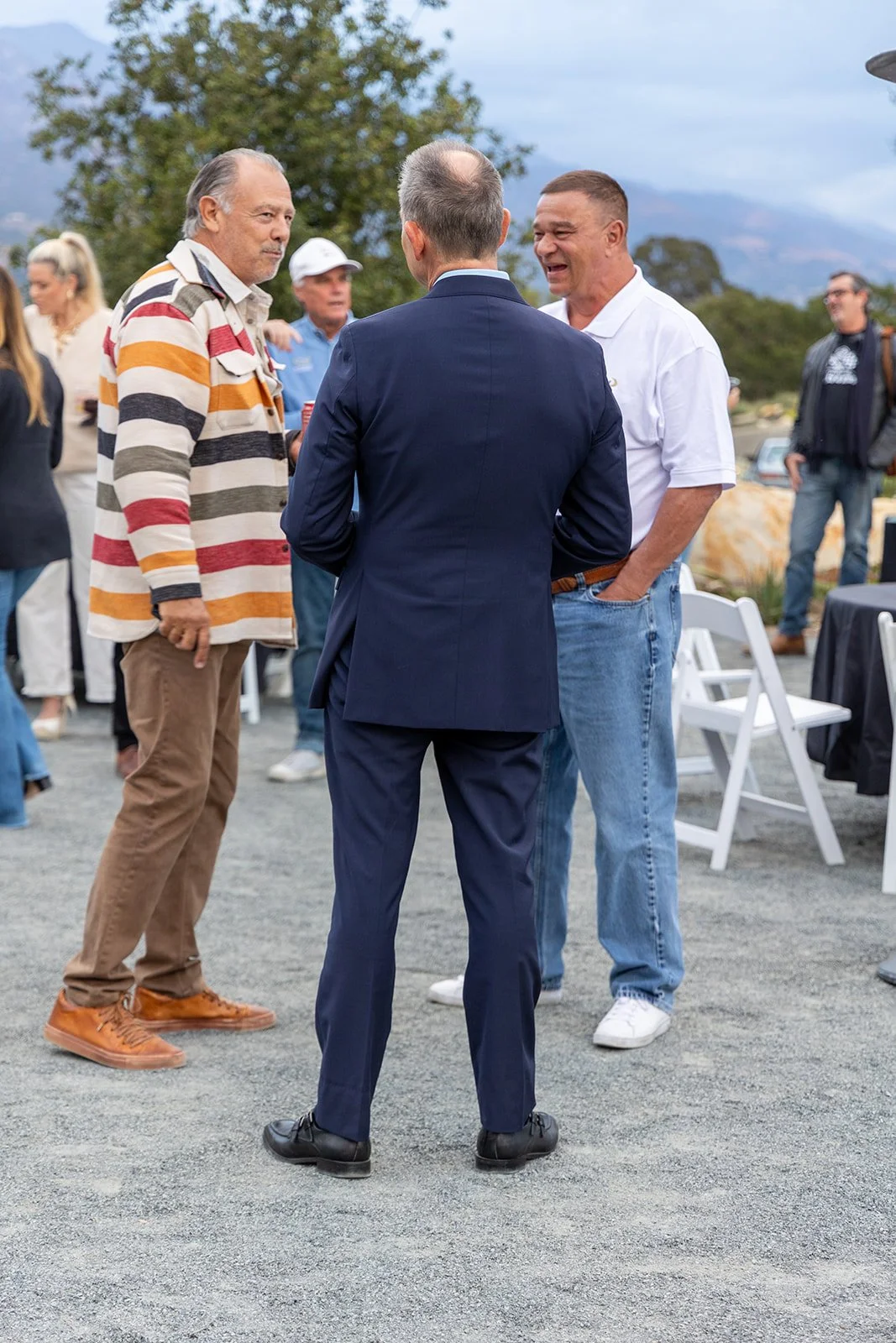 Group of people socializing outdoors, with three men in the foreground engaged in conversation, one wearing a striped shirt, another in a white polo shirt, and a third in a navy suit with his back to the camera. There are chairs and tables in the bac