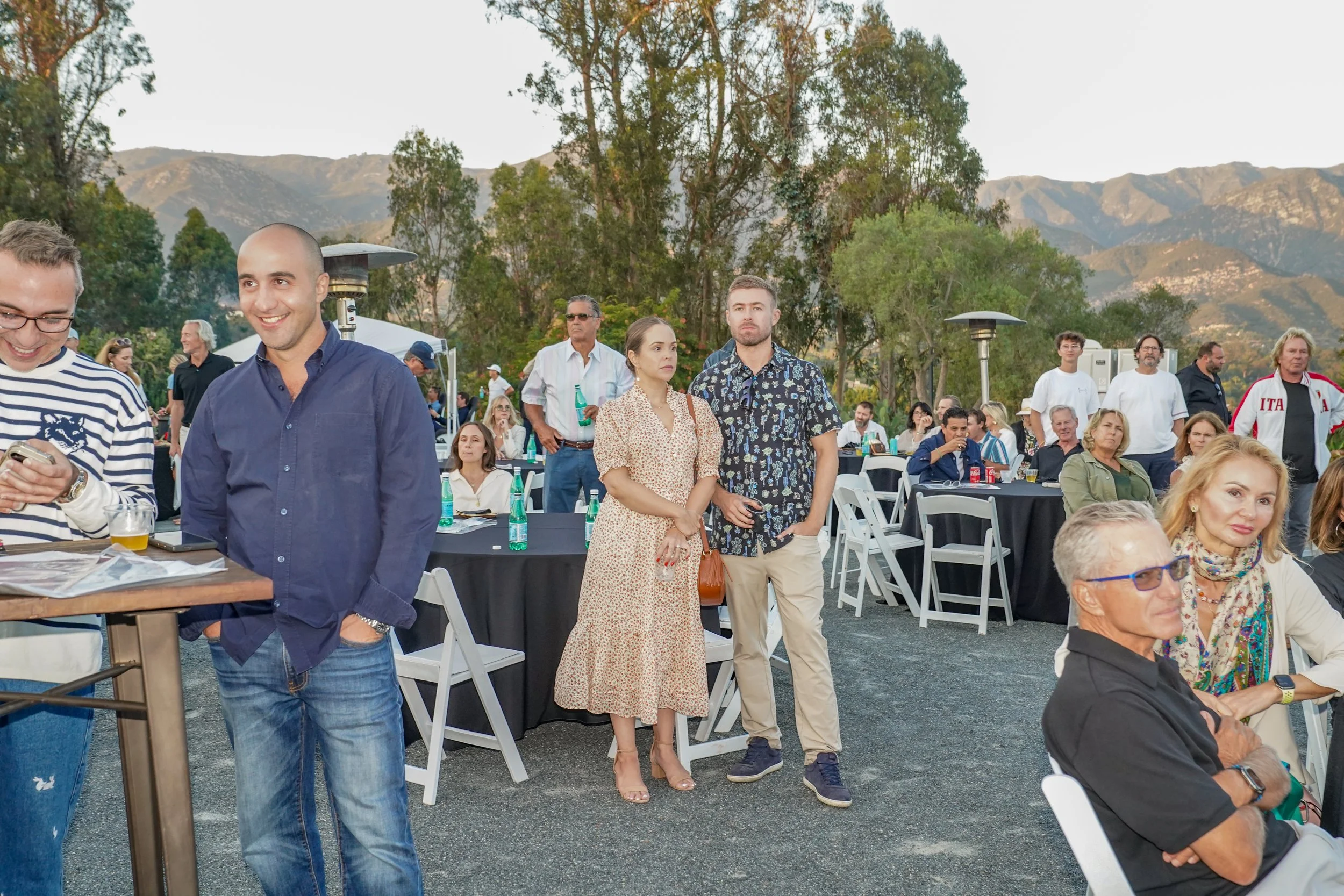 People gathered at an outdoor event with mountains in the background. Some are standing, others sitting at tables with drinks.