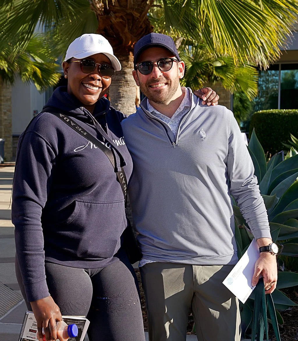 A smiling woman and man standing outdoors in a sunny area with palm trees, dressed casually with hats and sunglasses. They are close together, with the woman holding a notebook and a water bottle, and the man holding papers and a tote bag.