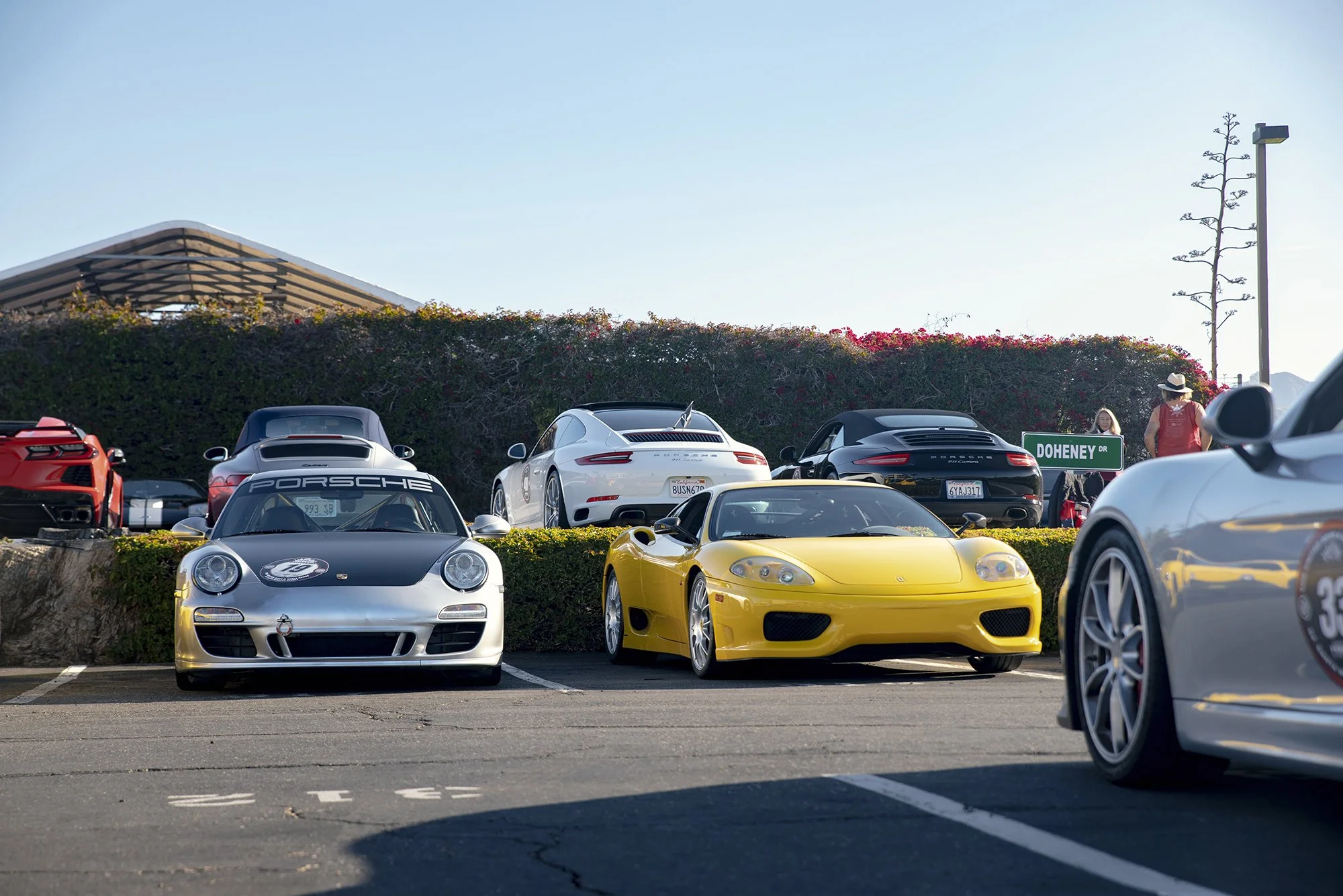 A parking lot featuring several luxury sports cars, including a yellow Ferrari, a silver Porsche, and a red car, with a hedge, a blue sky, and people walking nearby.