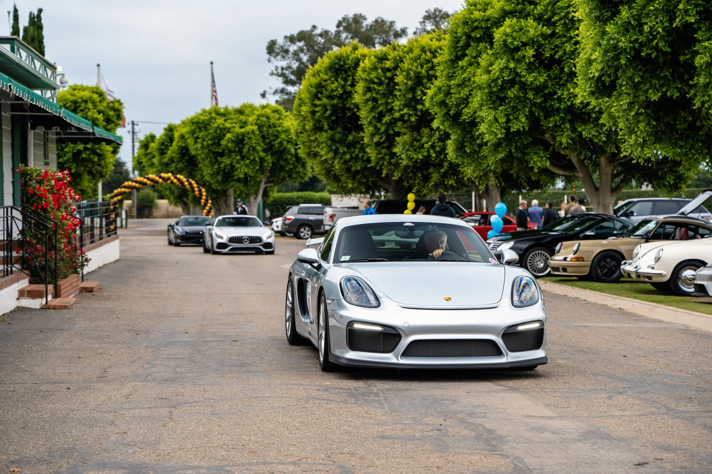 Silver sports car driving down a street lined with classic and luxury cars, with people and trees in the background.