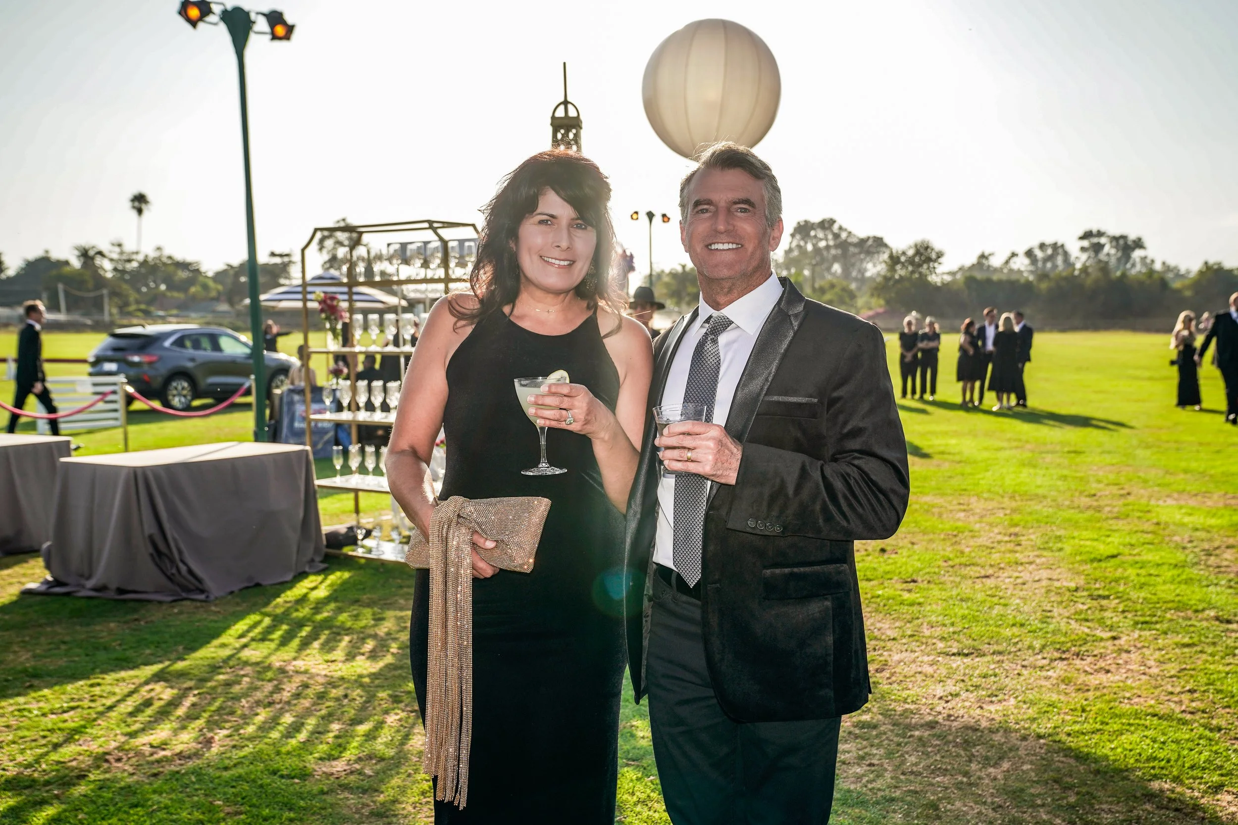 Man and woman in formal attire at an outdoor event, smiling and holding drinks, with a grassy area, decorations, and other guests in the background.