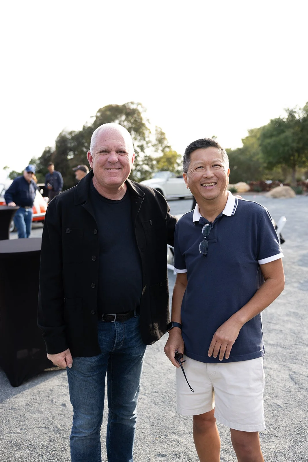 Two smiling men standing outside, one wearing a black jacket and jeans, the other wearing a navy polo shirt and white shorts.