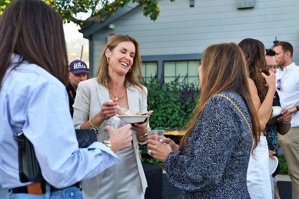 A group of people socializing outdoors, with some holding plates and drinks, and smiling while engaging in conversation.
