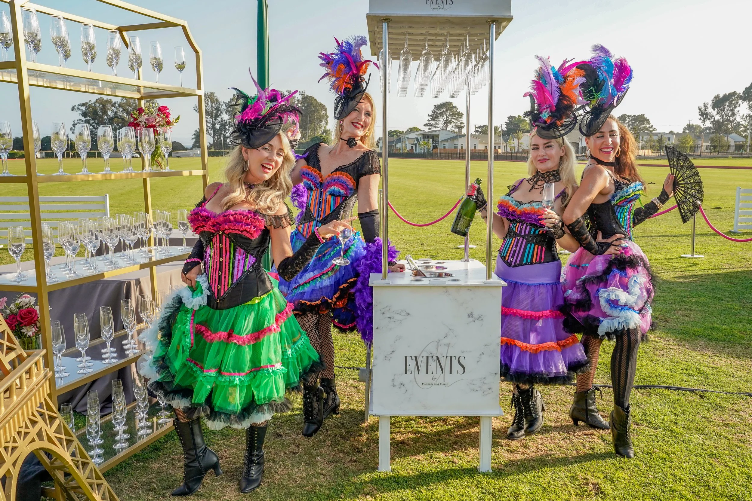Four women dressed in colorful, elaborate costumes with feathered hats, posing at a champagne event outdoors on a grassy field.