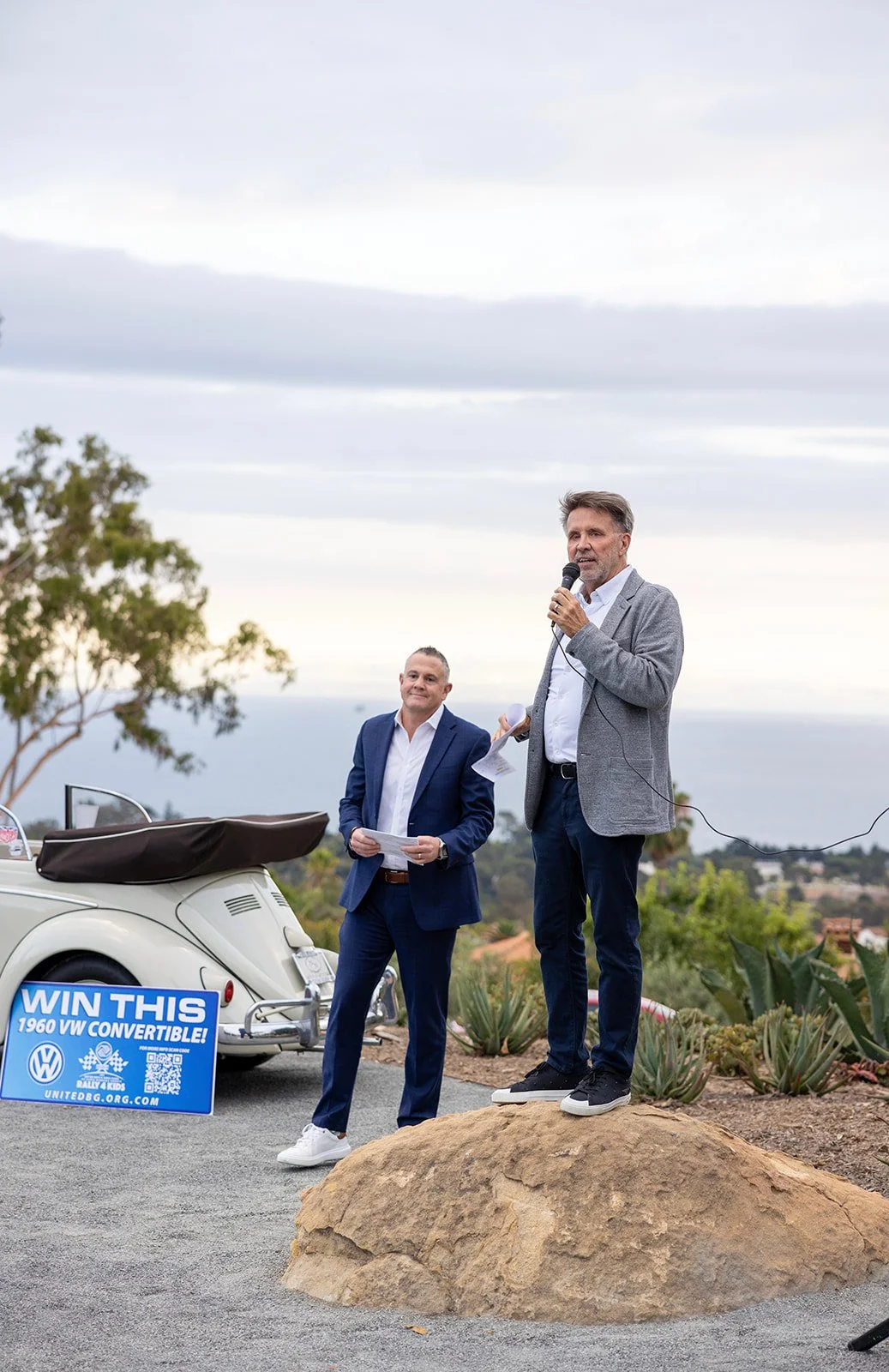 Two men standing outdoors, one speaking into a microphone and the other holding papers, with a vintage convertible car and a sign in the background.