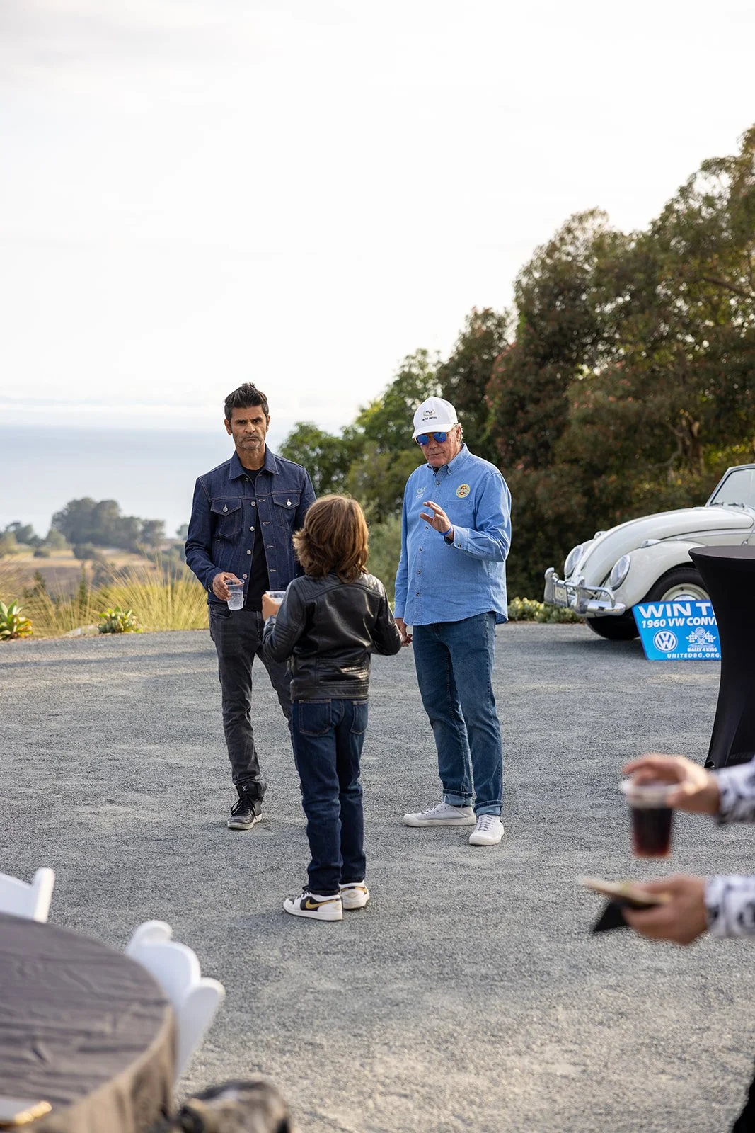 Three people are standing outdoors near a white vintage Volkswagen Beetle car. One man with dark hair in a denim jacket and dark pants is holding a clear cup, and another man with white hair, sunglasses, a white cap, and a blue shirt is talking to a 
