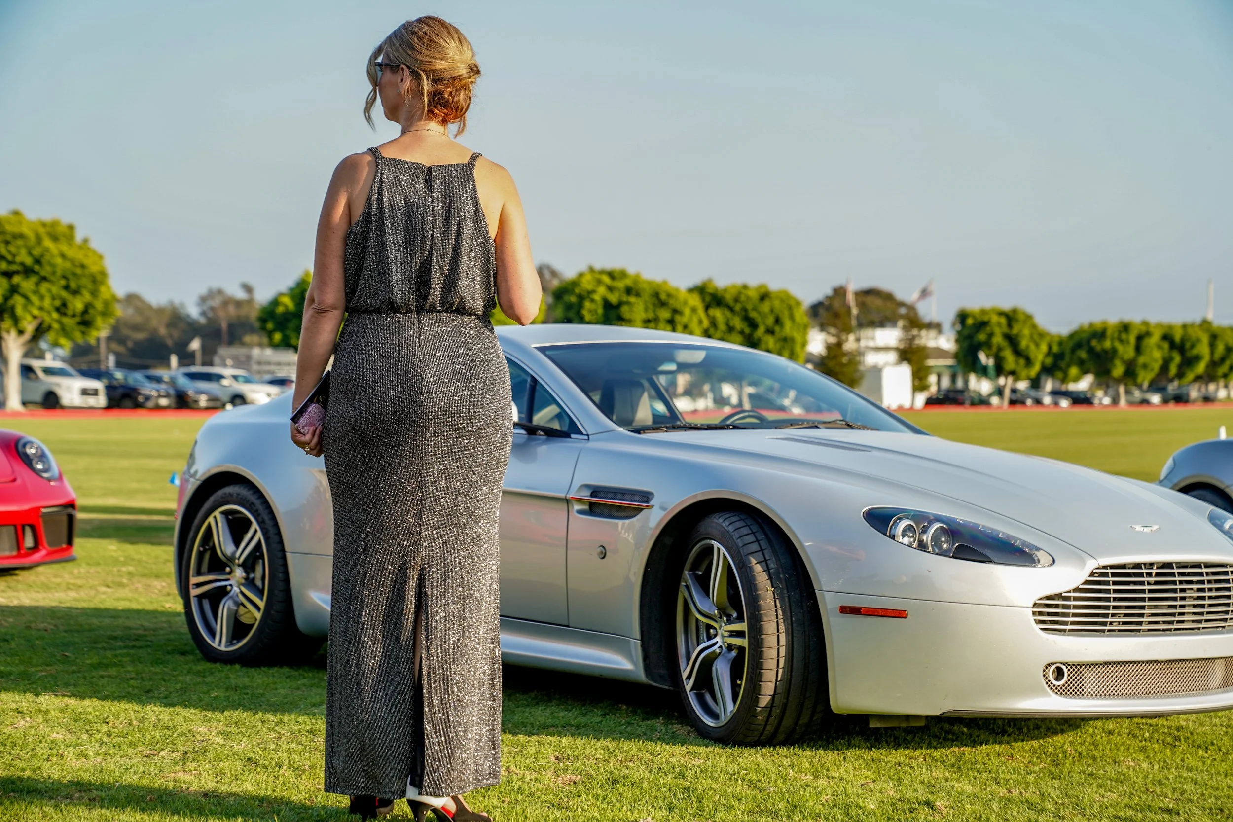 A woman in a sparkly black dress and sunglasses stands on grass, with a silver sports car behind her at an outdoor event, with trees and a parking lot in the background.