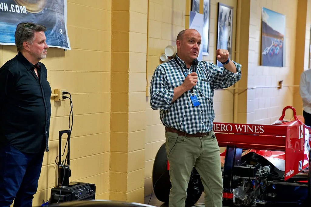 Two men standing indoors near a yellow brick wall, one speaking into a microphone, the other listening. There is a red race car with the words 'HERO WINES' on the rear wing behind them.