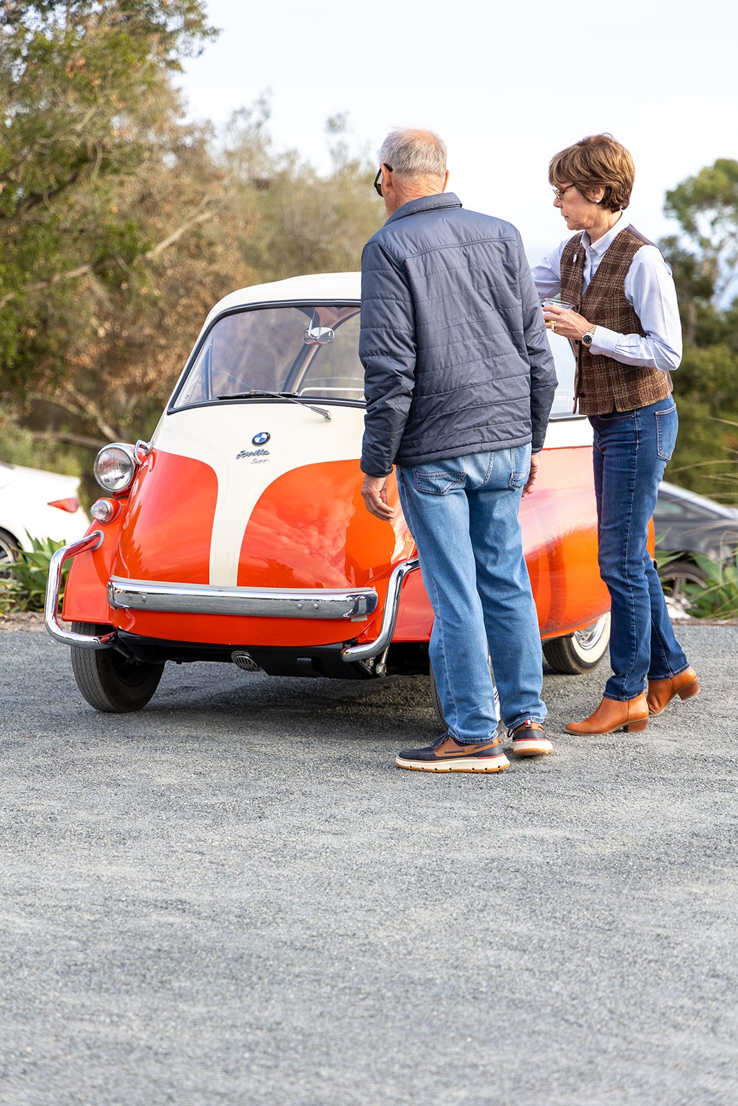 Two people talking next to a vintage BMW Isetta microcar outdoors, with trees and cars in the background.