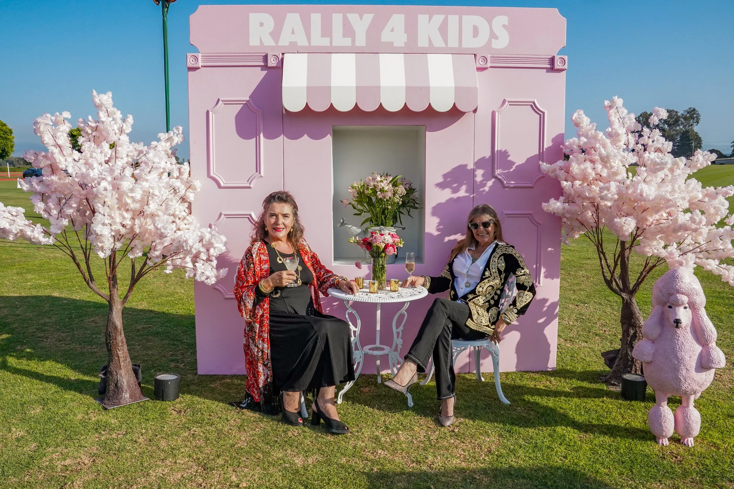 Two women sitting at a small round table with flowers, celebrating at a pink backdrop labeled 'RALLY 4 KIDS' with cherry blossom trees and pink poodle sculpture outdoors.