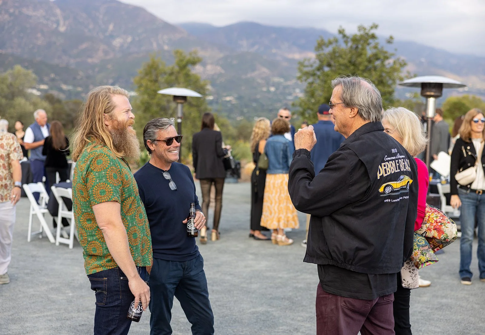 Group of people socializing outdoors, mountains in the background, several individuals holding drinks, engaging in conversation at an event.
