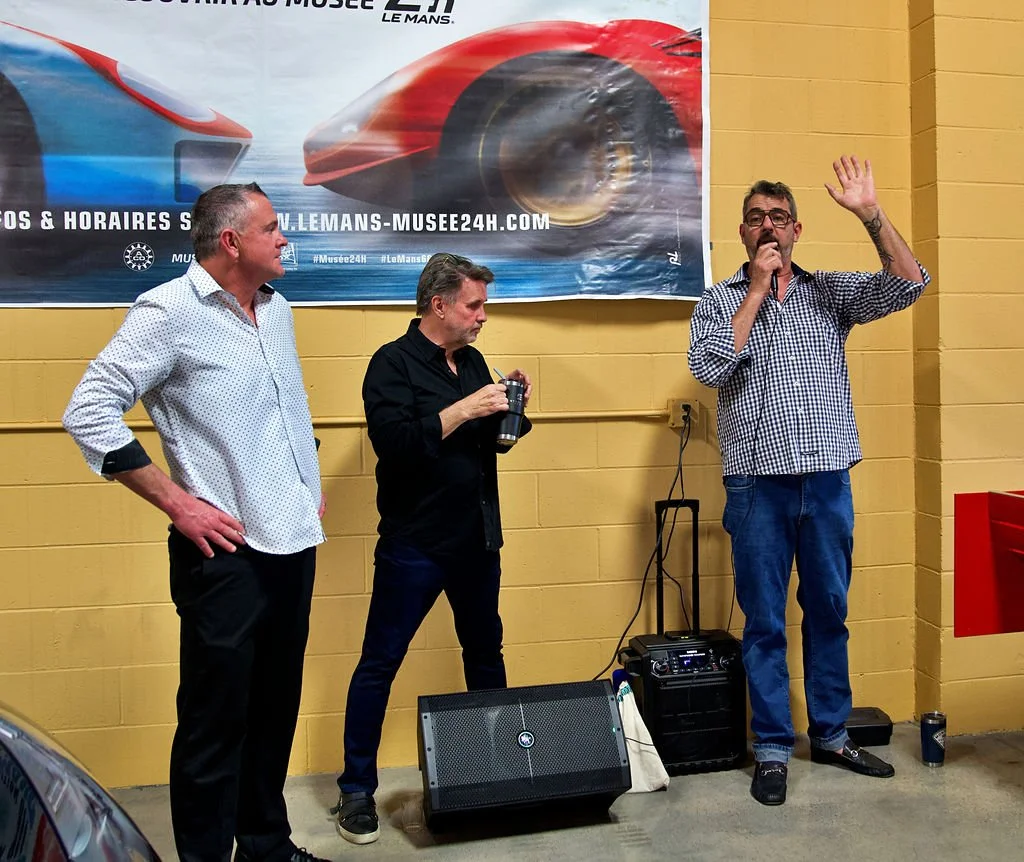 Three men standing in front of a large banner featuring race cars at an indoor event. One man is speaking into a microphone and waving, another is holding a cup, and the third is looking at the speaker.