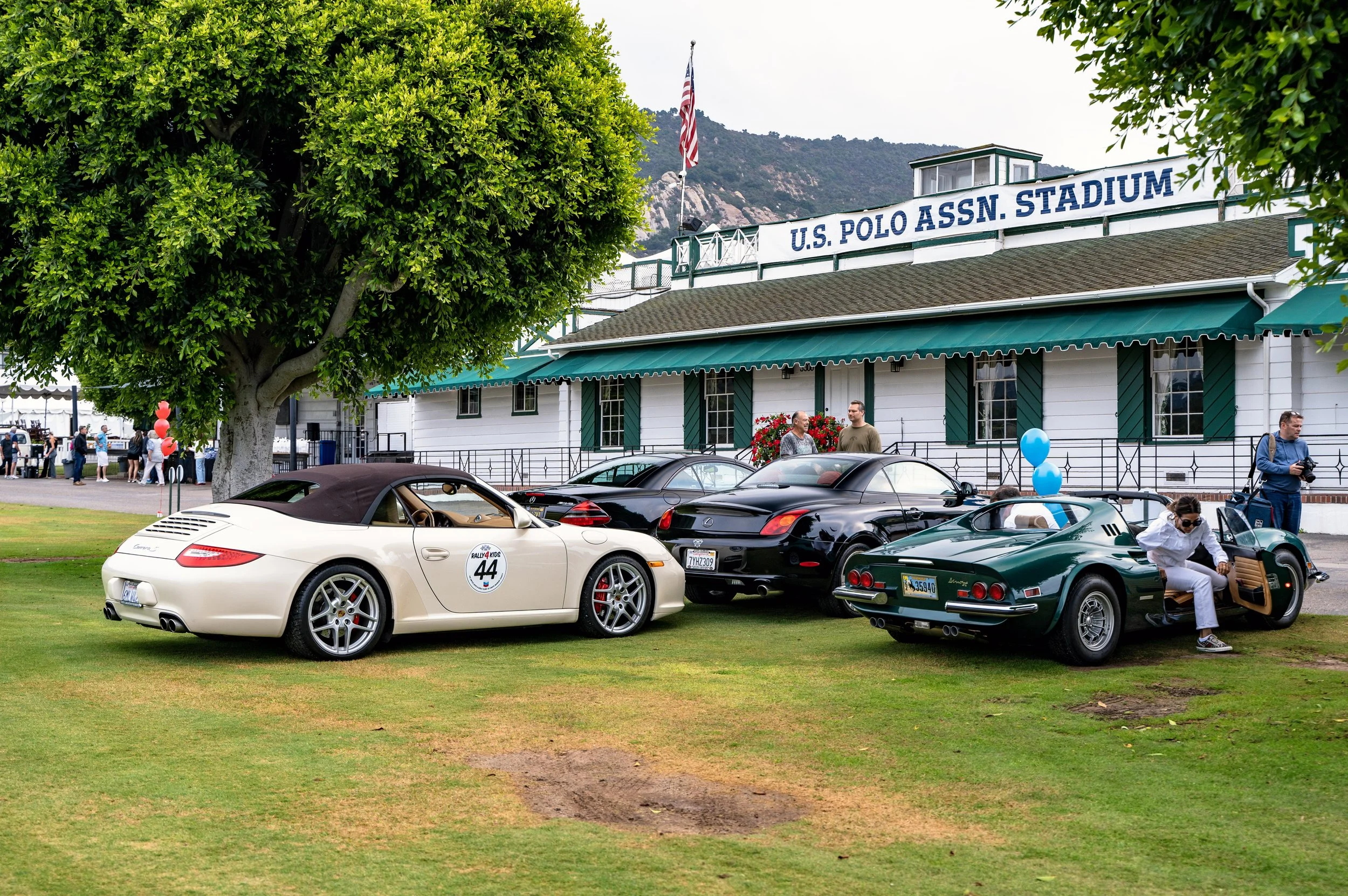 Classic and modern sports cars parked on the grass in front of the U.S. Polo Assn. Stadium during an outdoor car event, with people walking and talking in the background, and a large tree providing shade.