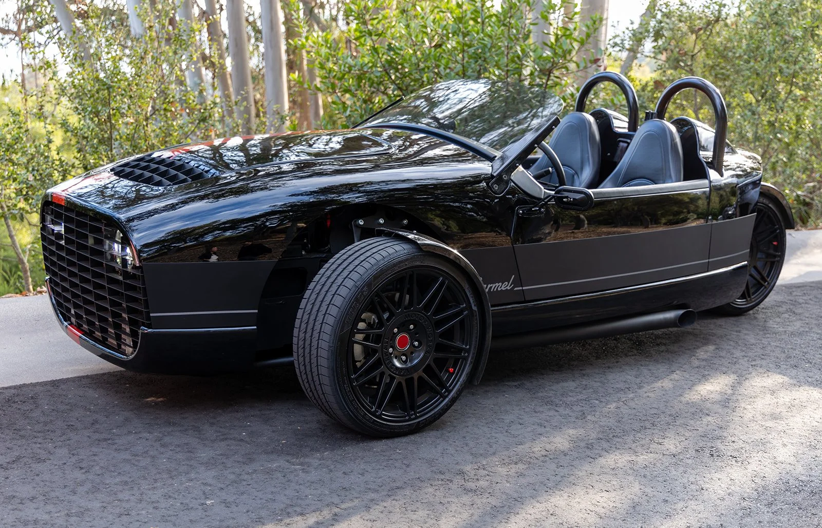A sleek black sports car with an open top parked on the side of a road surrounded by trees.