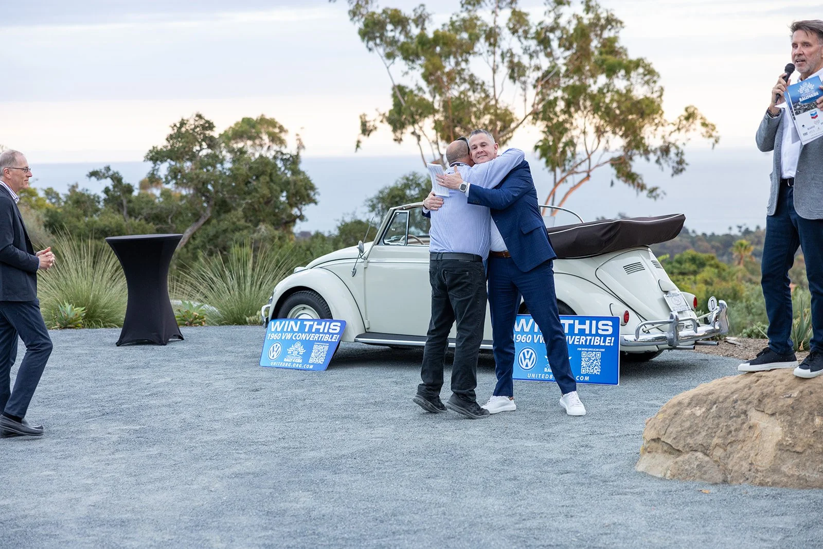 Two men hugging in front of a vintage white Volkswagen convertible at an outdoor event, with signs indicating a contest to win the car and a man speaking on a microphone on the right.