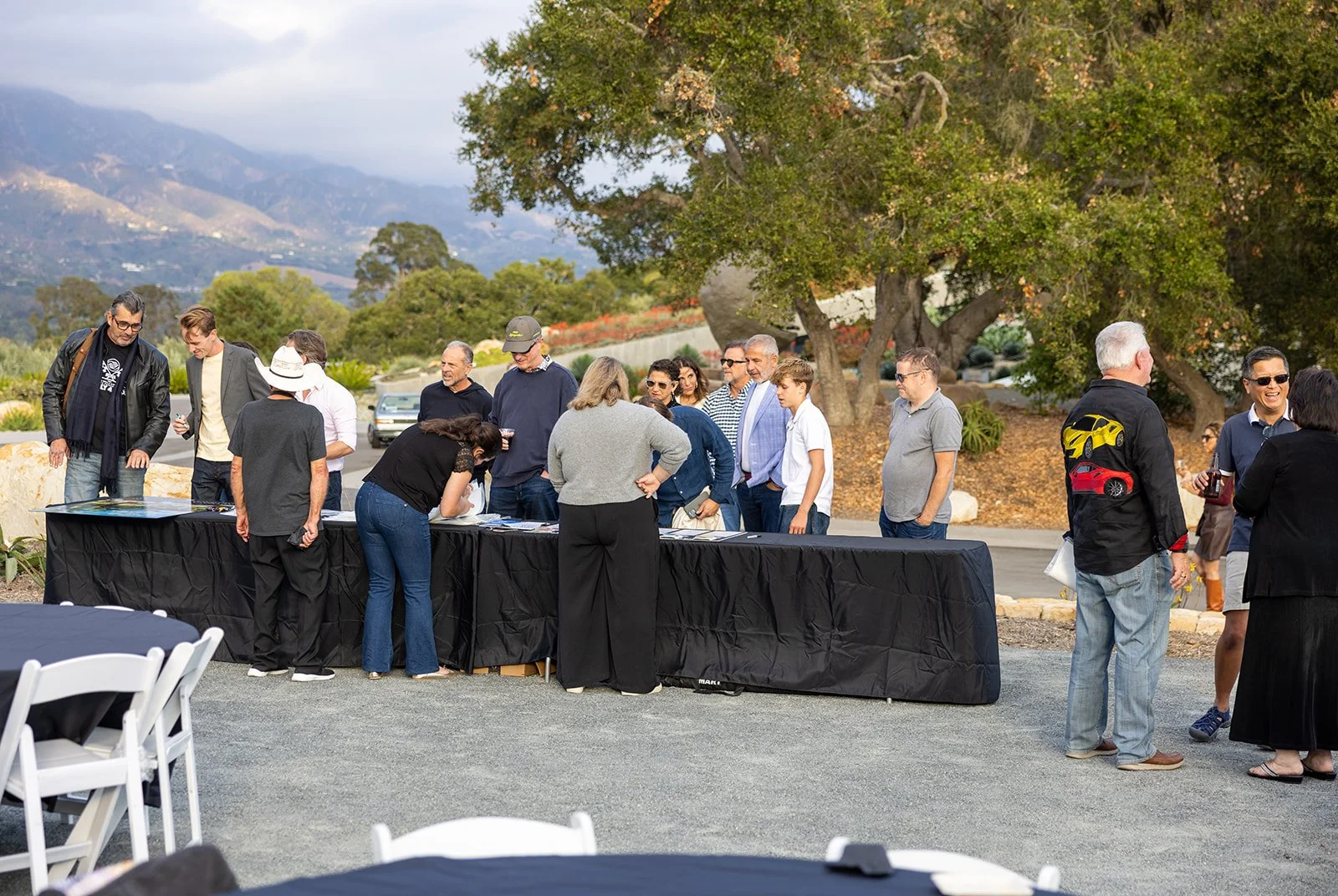 A group of people gathered outdoors around a long black table, engaging with documents or materials, with mountains and trees in the background.