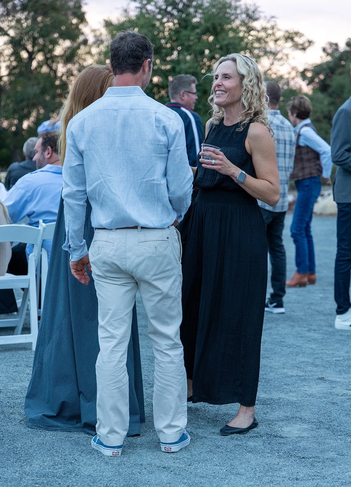 A woman with curly blonde hair wearing a black sleeveless dress and a smartwatch, smiling and holding a drink, converses with a man in a light blue shirt and white pants at an outdoor social gathering during evening. Several other people are in the b