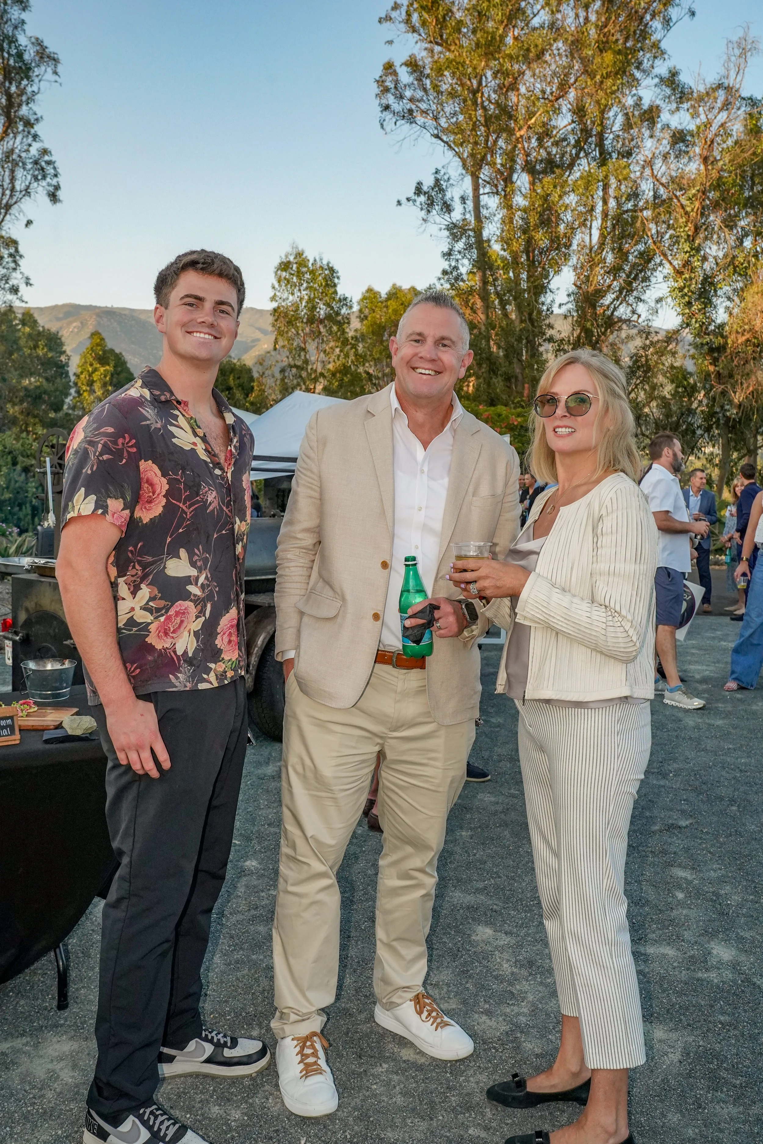 Three people standing outdoors at a social event, smiling at the camera. Two men and one woman, dressed in casual and business casual attire, with trees and a mountain in the background.