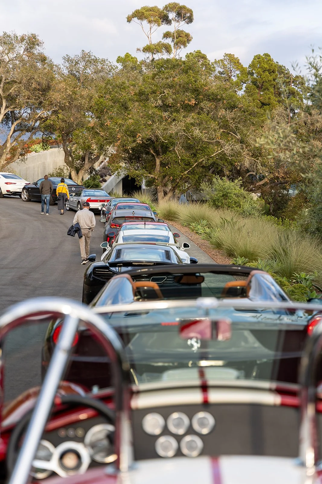 Line of luxury sports cars parked on a scenic hillside, viewed from the drivers' seats of a vintage race car.