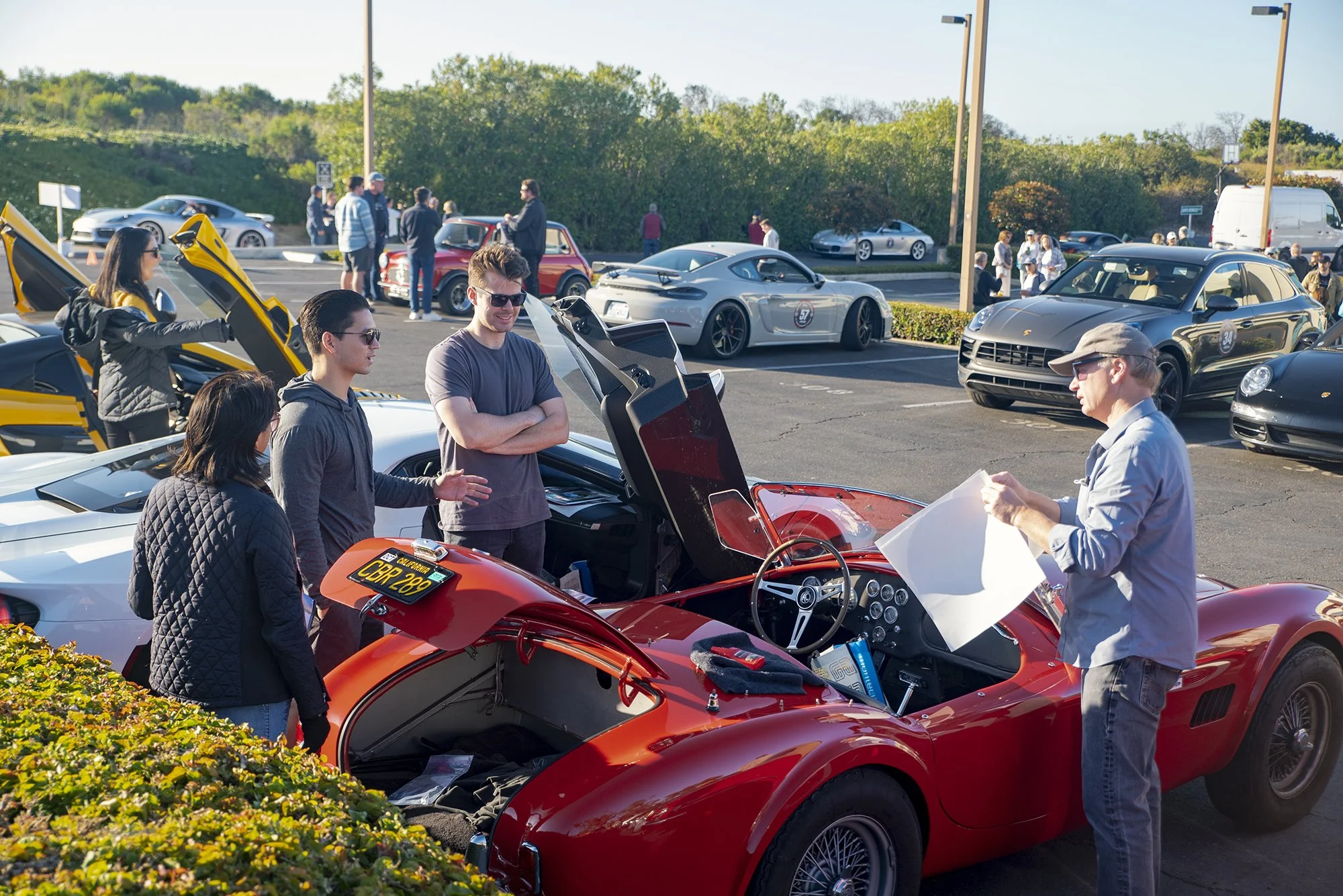 People gathered around a red vintage sports car at a car show, with other luxury and sports cars parked in the background on a sunny day.