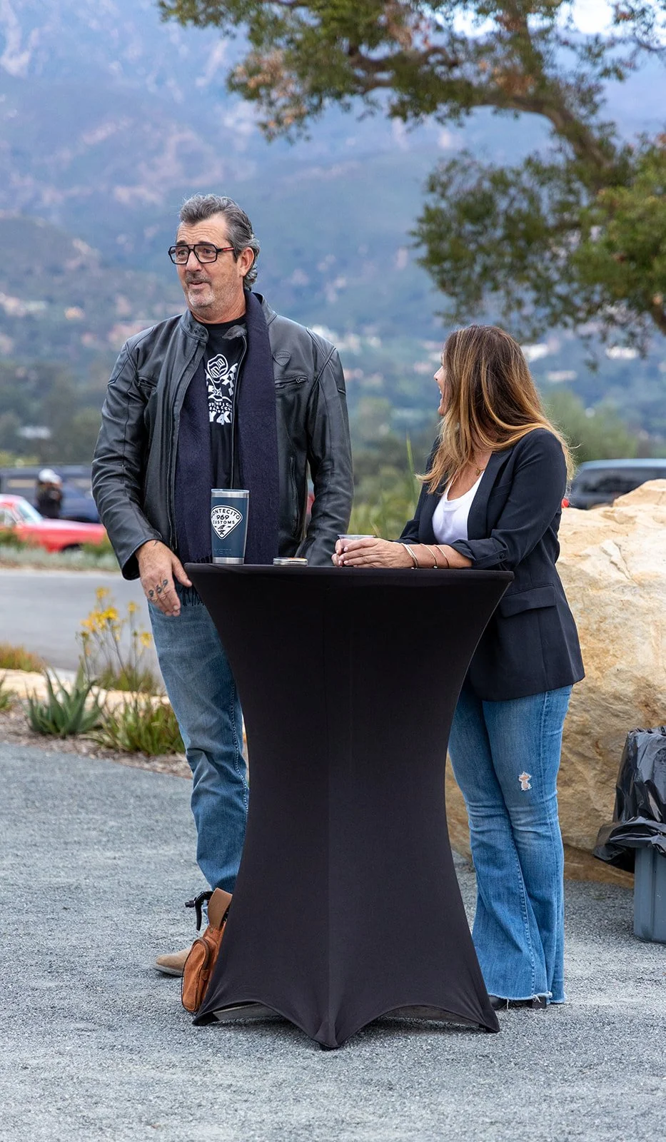 A man and a woman having a conversation outdoors near a table with drinks, with mountains and trees in the background.